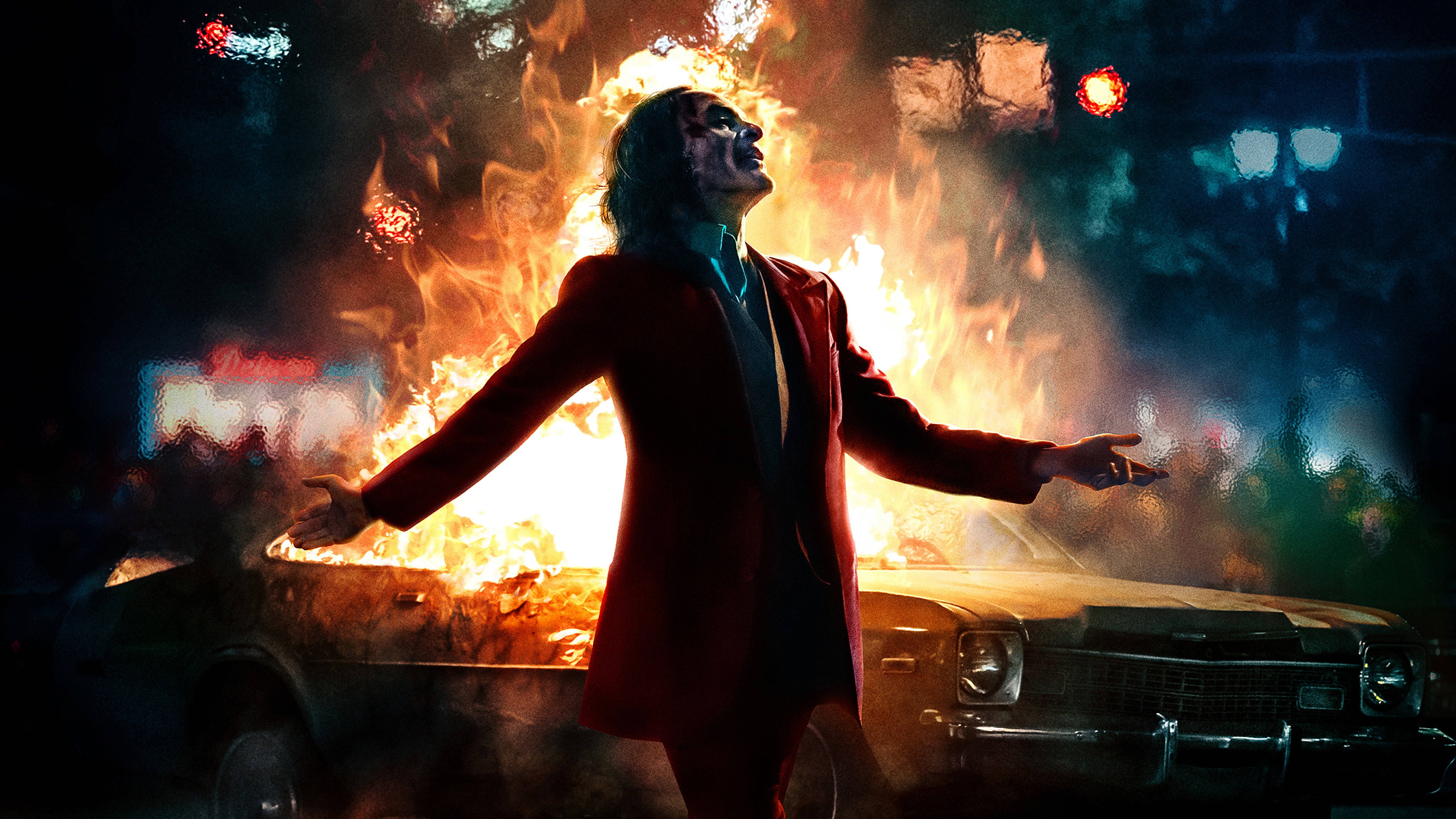 小丑 Joaquin Phoenix 桌面 4K 壁紙 — 一名身穿紅色西裝的男子在夜間霓虹燈下，張開雙臂站在一輛燃燒的汽車前。
