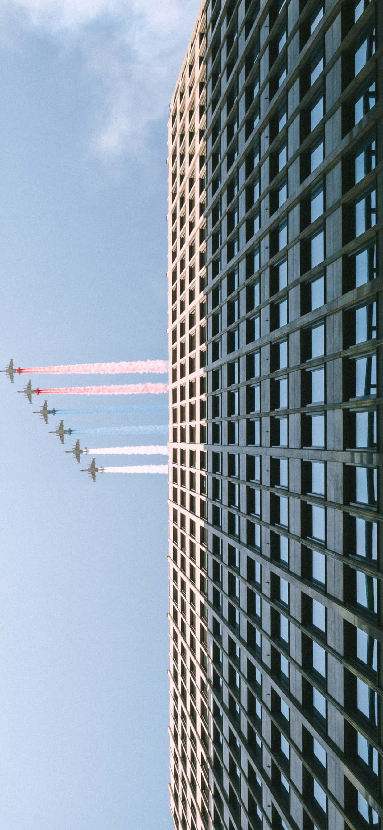 小型商用飛機飛越夕陽天空 壁紙 — 低角度拍攝戰鬥機編隊飛行，拖曳著彩色煙霧，掠過高聳的摩天大樓，背景是清澈的藍天。