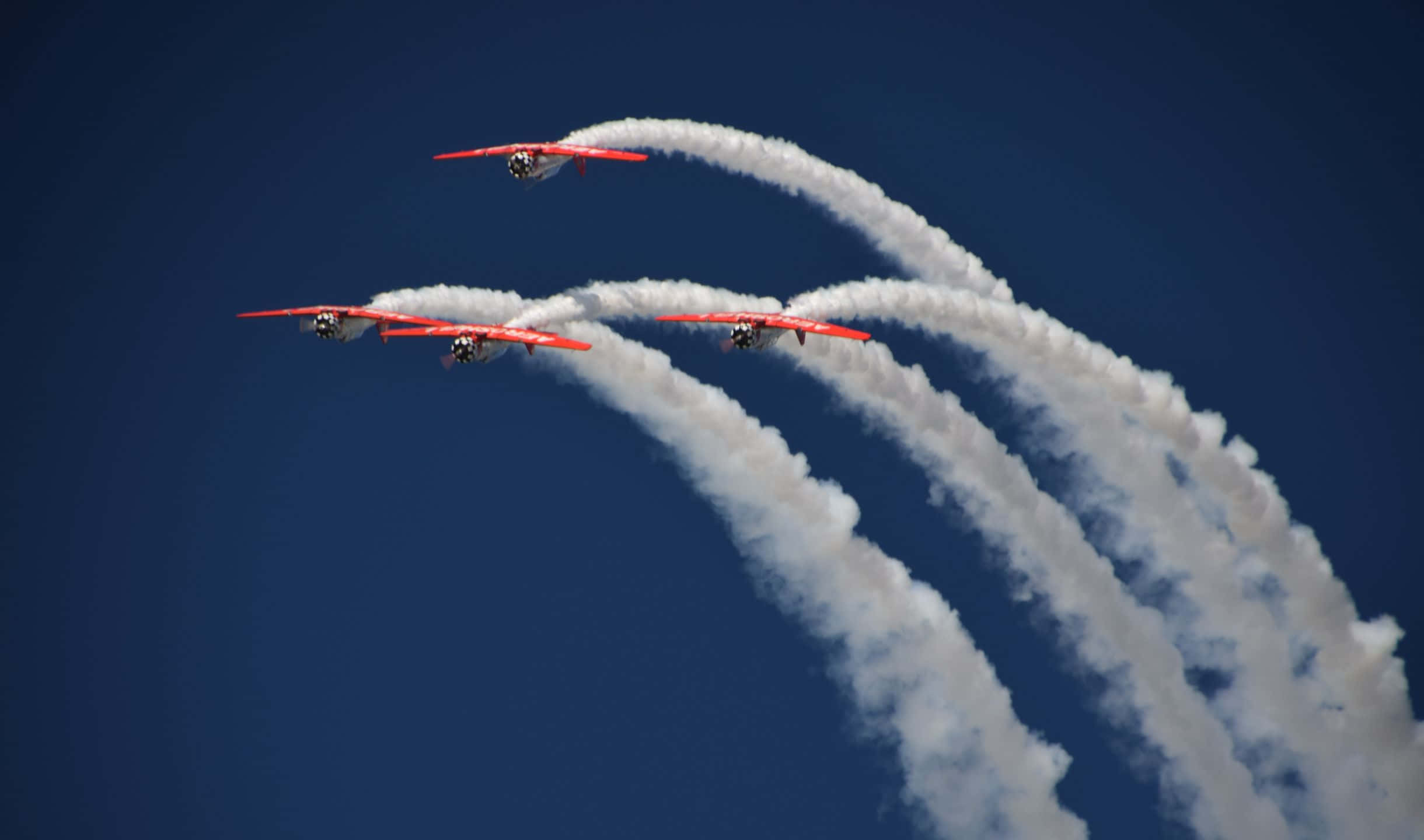 小型飛機編隊飛行的空中視角 壁紙 — 三架紅色雙翼機以緊密編隊翱翔於深藍色天空，留下濃厚的白色煙霧軌跡。