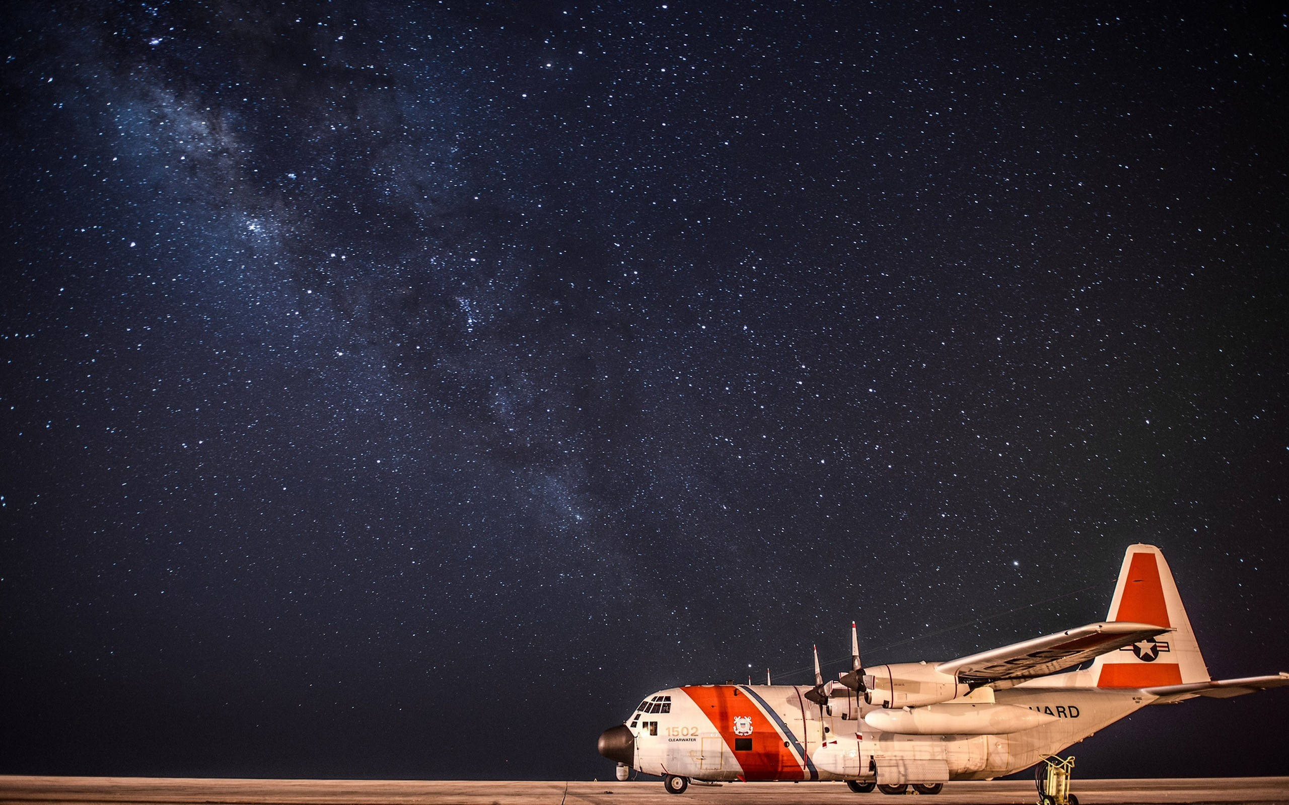 小型飛機與星系 壁紙 — 一架海岸防衛隊飛機停在跑道上，上方是令人驚嘆的星空，銀河拱起。