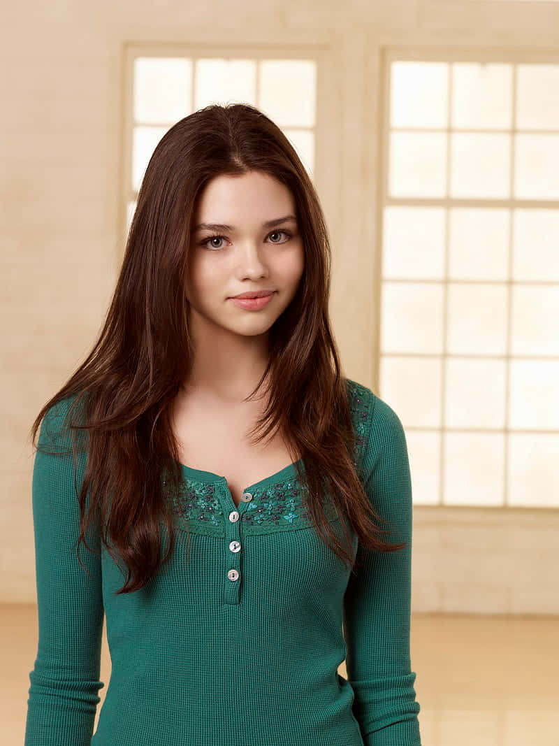 小巧的India Eisley 壁紙 — 一位長棕髮的年輕女性，在光線柔和、有大窗戶的房間裡穿著一件青綠色鈕扣上衣。