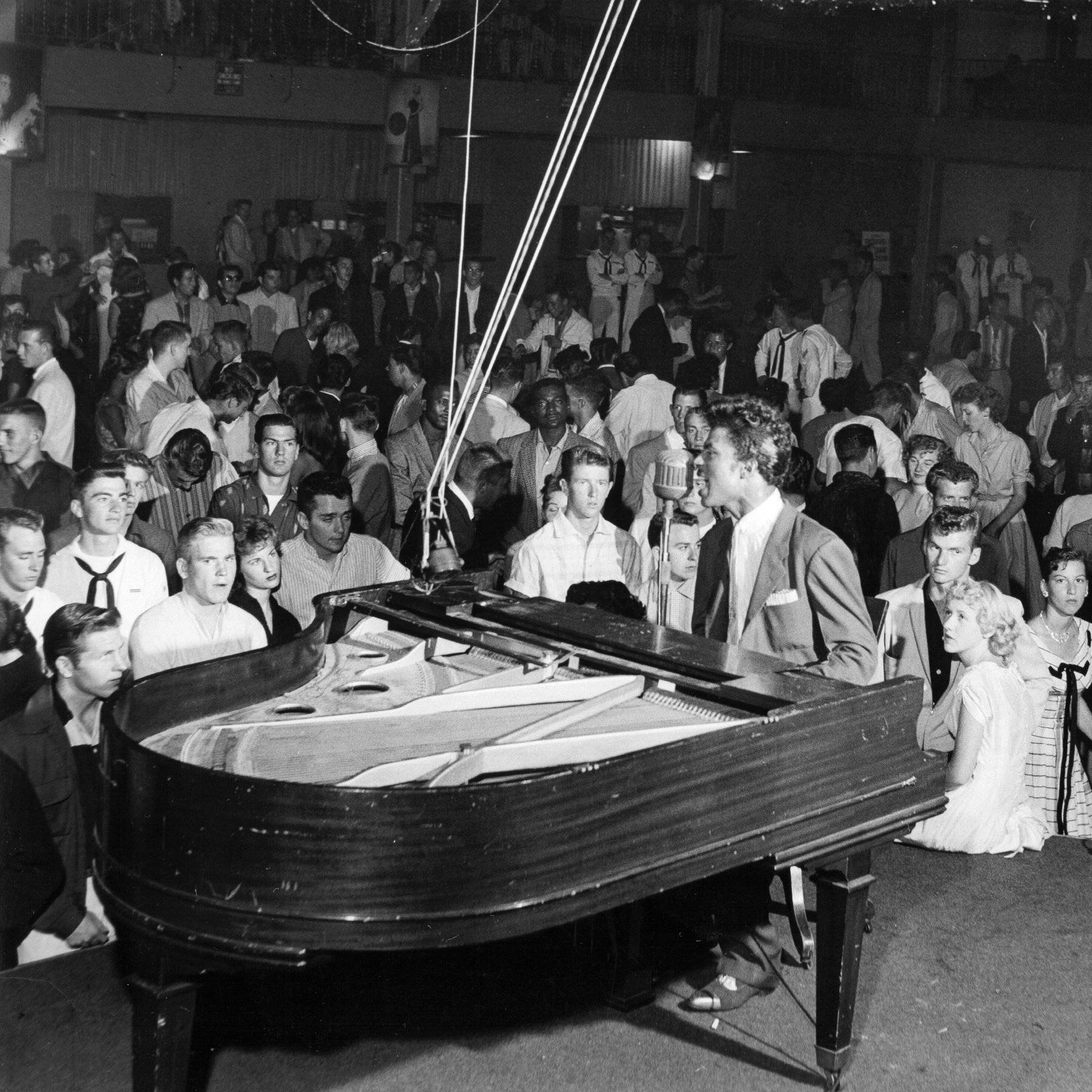 小理查德1955年舞台鋼琴演奏現場 壁紙 — 一張復古黑白照片捕捉到一位男歌手在昏暗的場地中，於滿座的現場觀眾前在三角鋼琴上表演。
