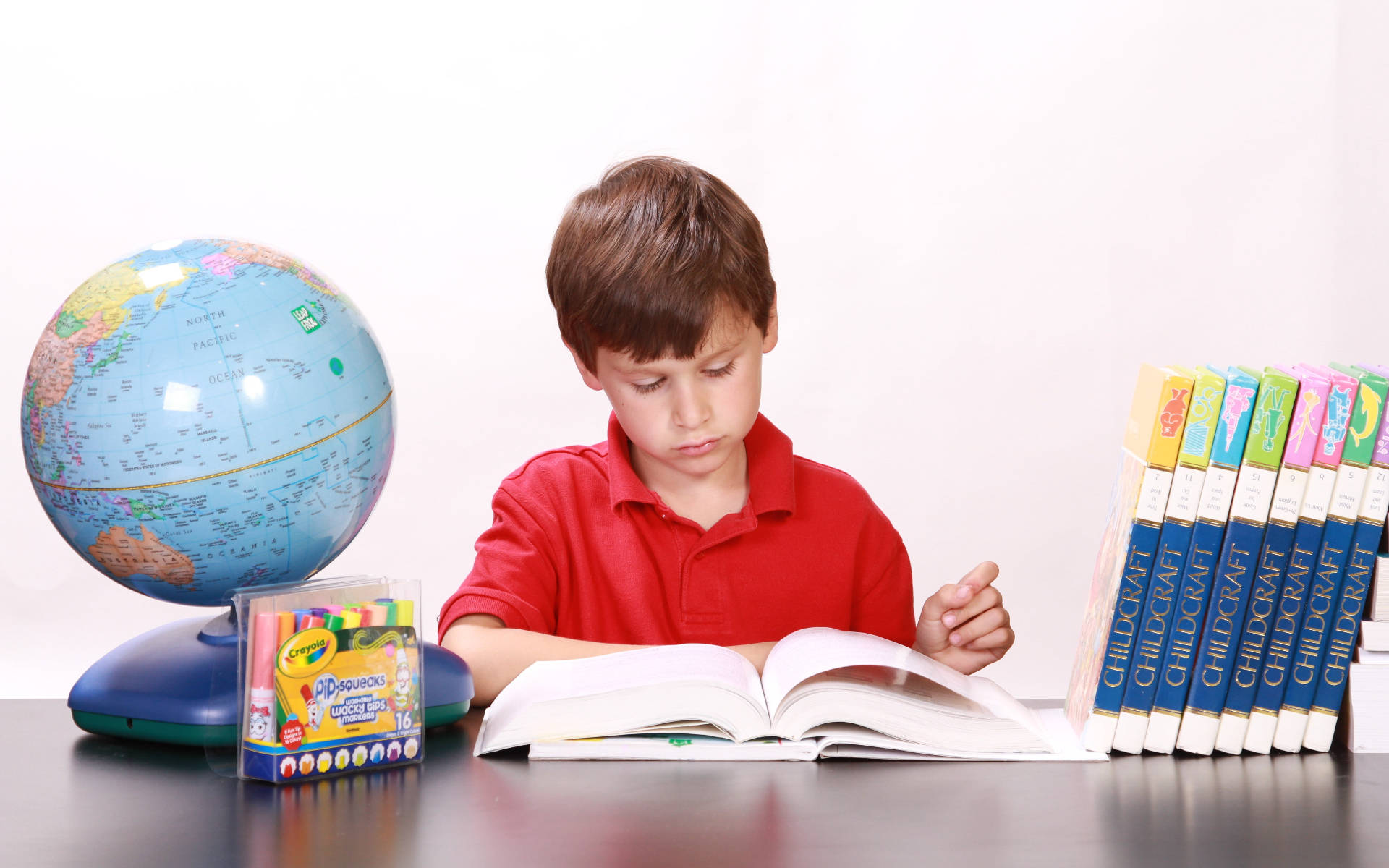 小男孩閱讀教育書籍 壁紙 — 一個穿著紅色襯衫的小男孩在書桌前看書，周圍有地球儀、蠟筆和一堆Childcraft書籍。