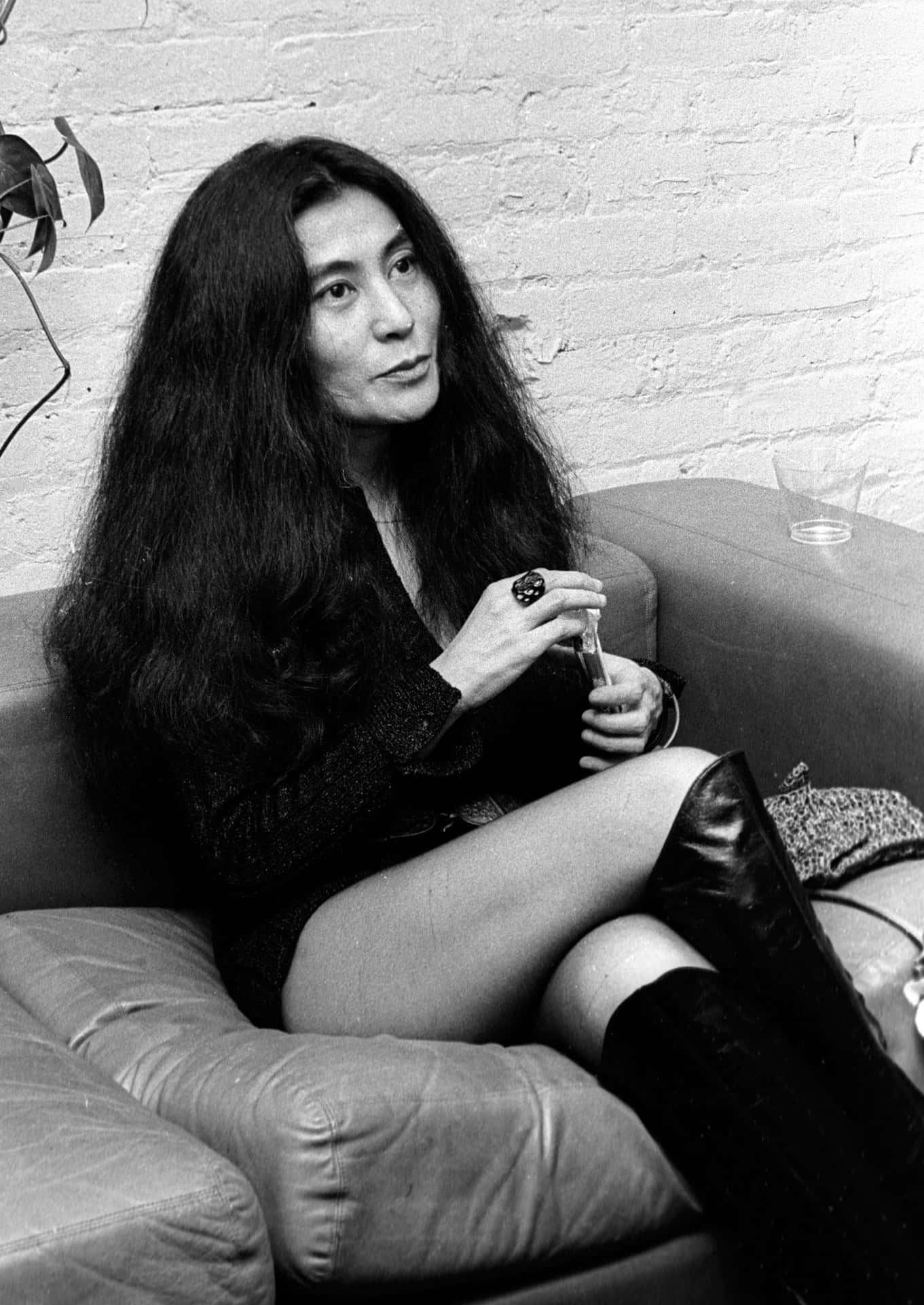 長黑髮女性坐在皮革沙發上，手持玻璃杯，對著磚牆。