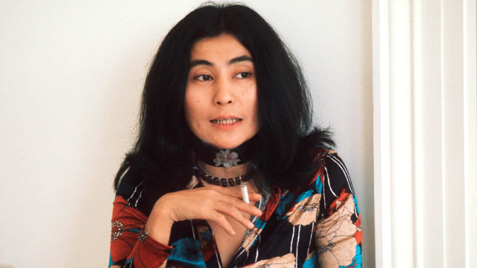 一位長黑髮的女性在室內手持香煙，對著鏡頭微笑。