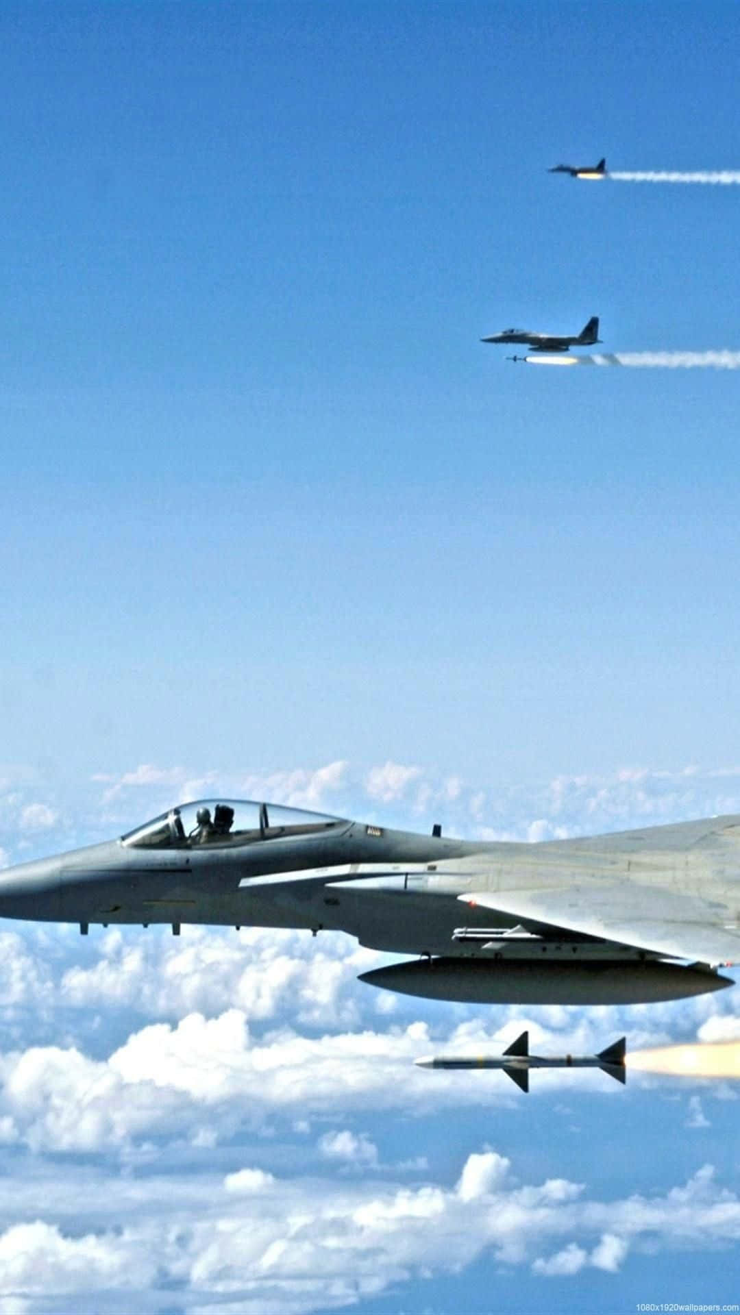 小飛機征服天空的壯麗畫面 壁紙 — 兩架戰鬥機在清澈的藍天中翱翔，飛越蓬鬆的白雲，其中一架發射導彈，留下煙霧痕跡。