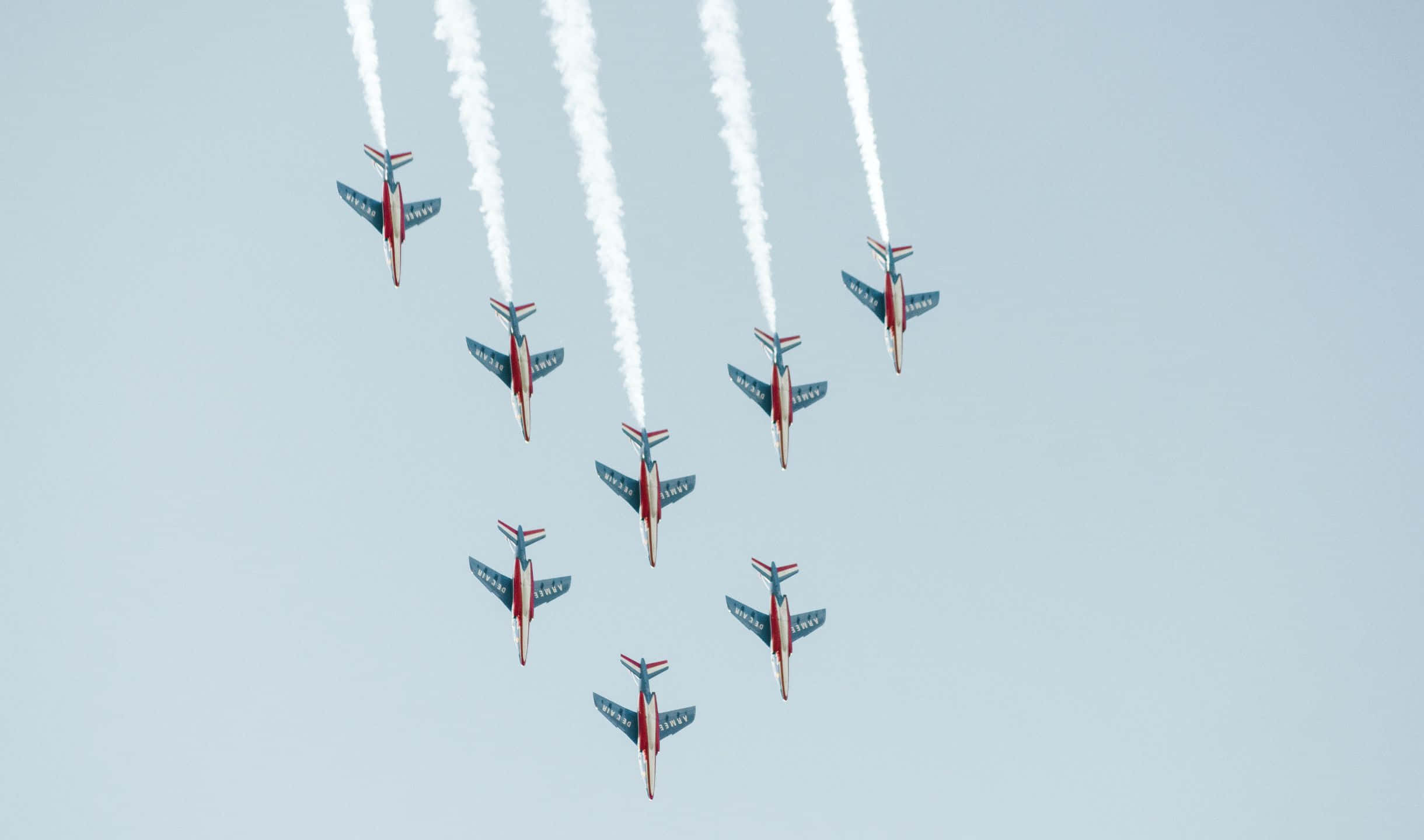 小飛機的空中視角 壁紙 — 七架紅、白、藍色噴射機以緊密編隊飛行，在淡藍色天空中留下濃厚的白色煙霧軌跡。