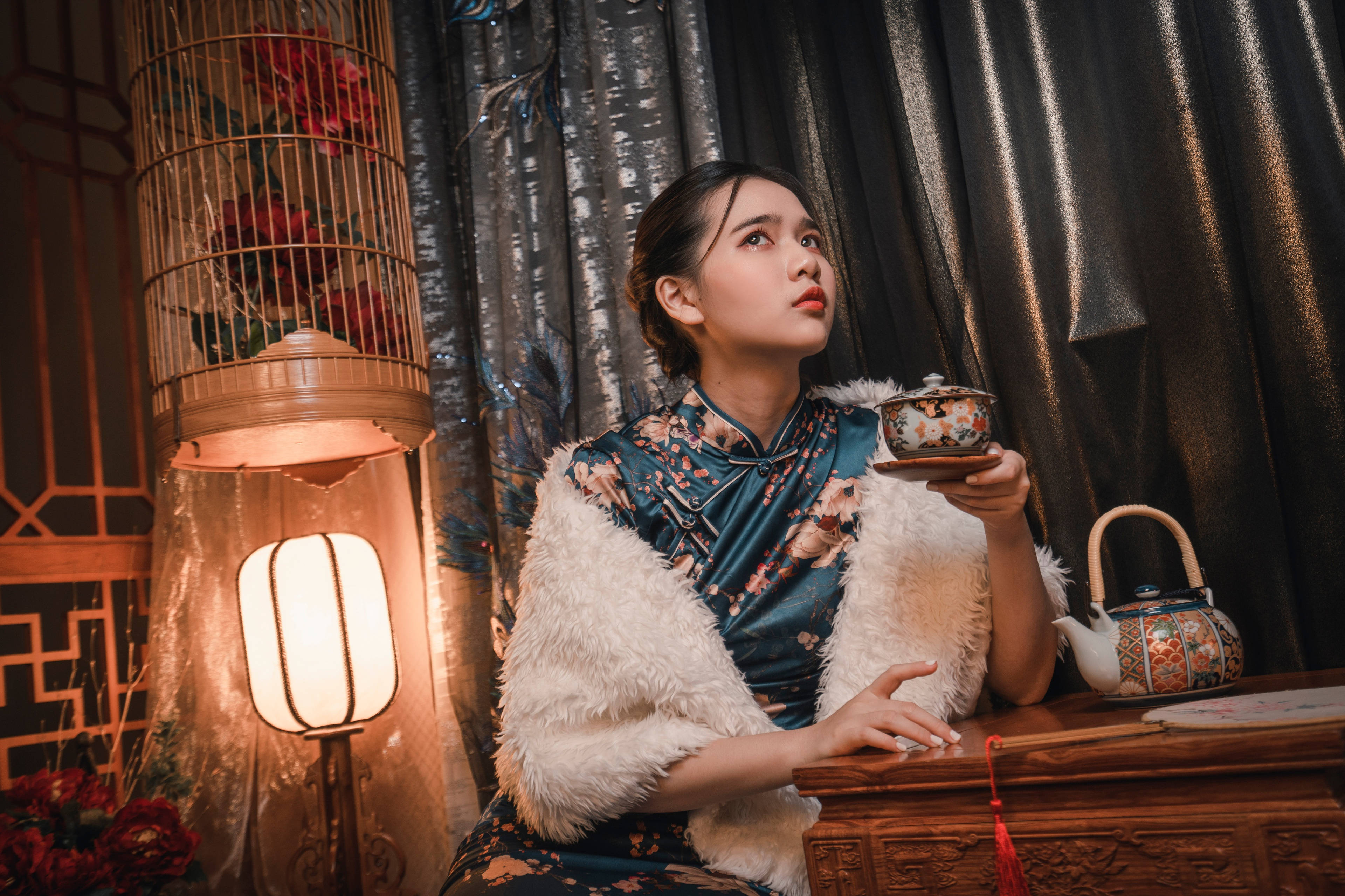 少女旗袍裝 壁紙 — 一位身穿花卉旗袍和白色毛皮披肩的女性，在溫暖的燈籠光下，拿著茶杯，旁邊是一個華麗的茶壺。