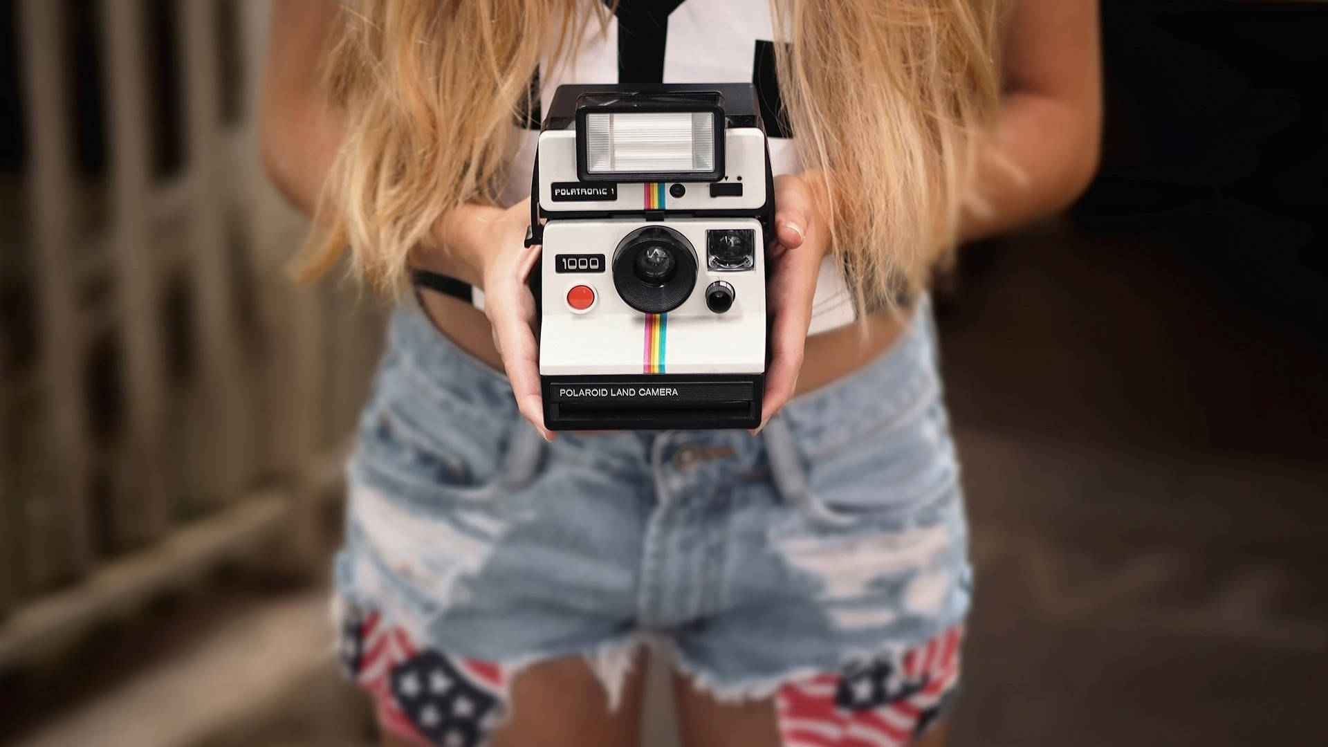 手持Polaroid Land Camera，身穿帶有美國國旗補丁的牛仔短褲。