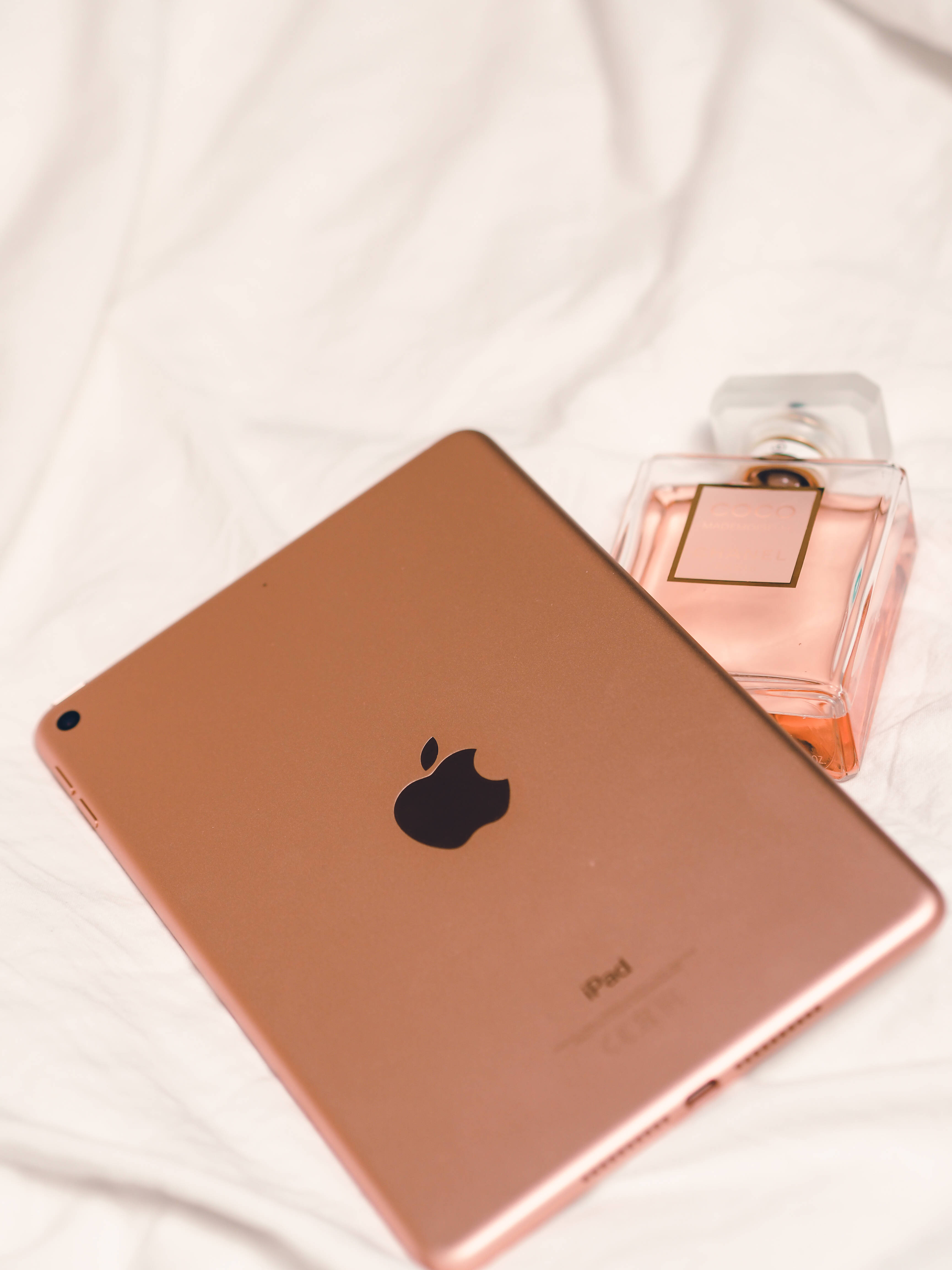 少女風手機電子產品與香水 壁紙 — 玫瑰金色iPad放在柔軟的白色布料上，旁邊放著一個相配的香水瓶。