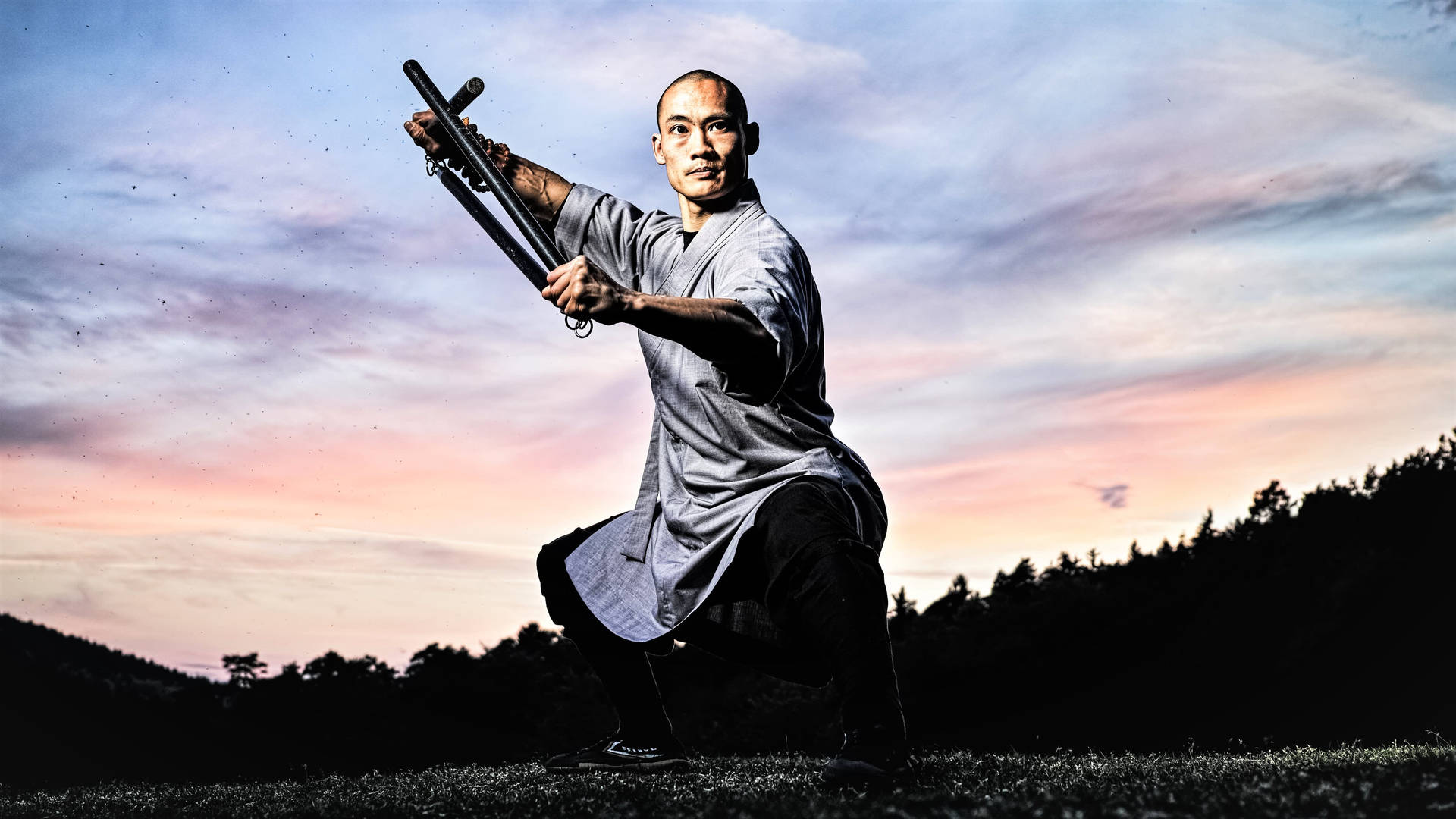 少林功夫總教練施恒毅 壁紙 — 一位身穿灰色長袍的武術家，在充滿活力的暮色天空下，用雙節棍擺出強而有力的姿勢。