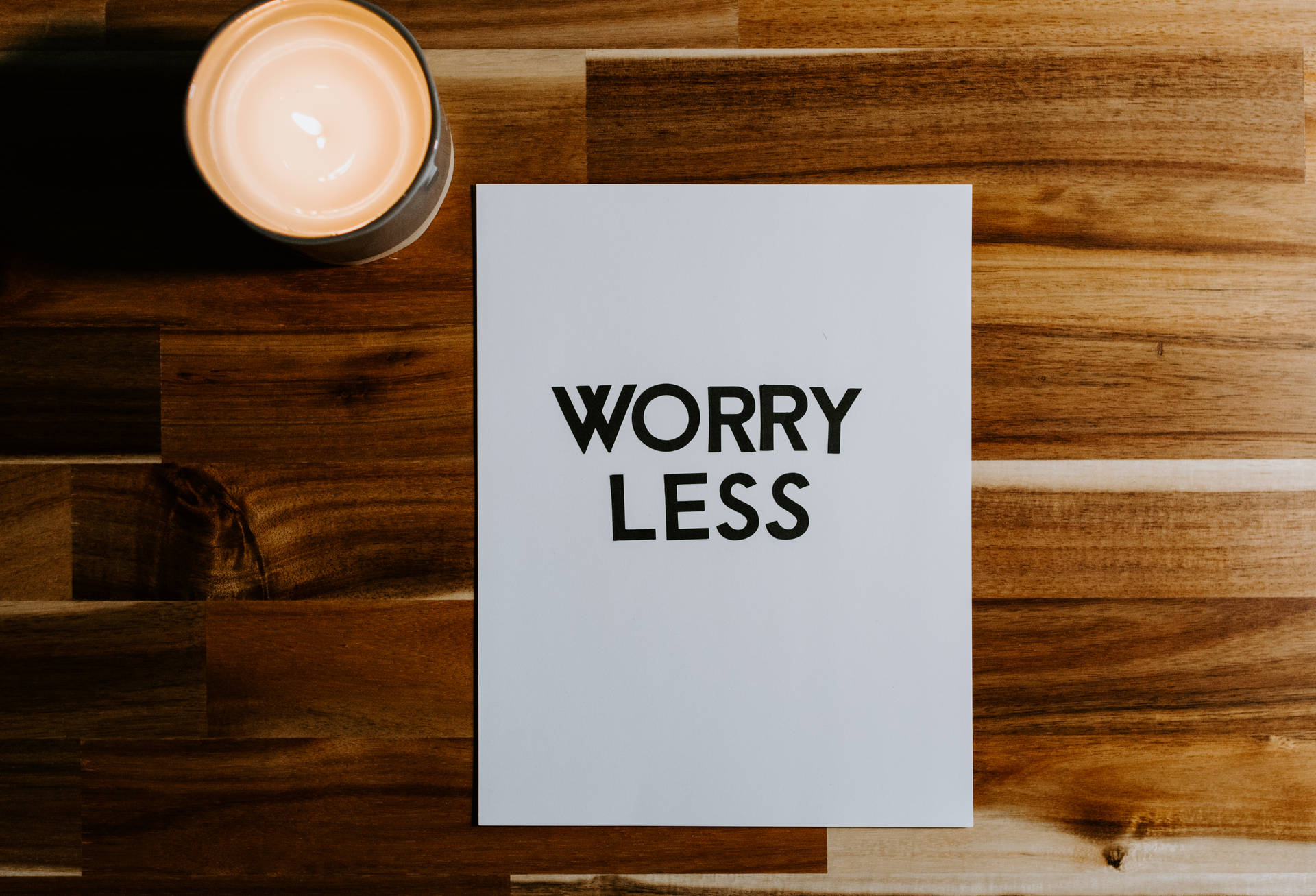 一支點燃的蠟燭旁邊放著一張寫有「WORRY LESS」的卡片，置於木質表面上。