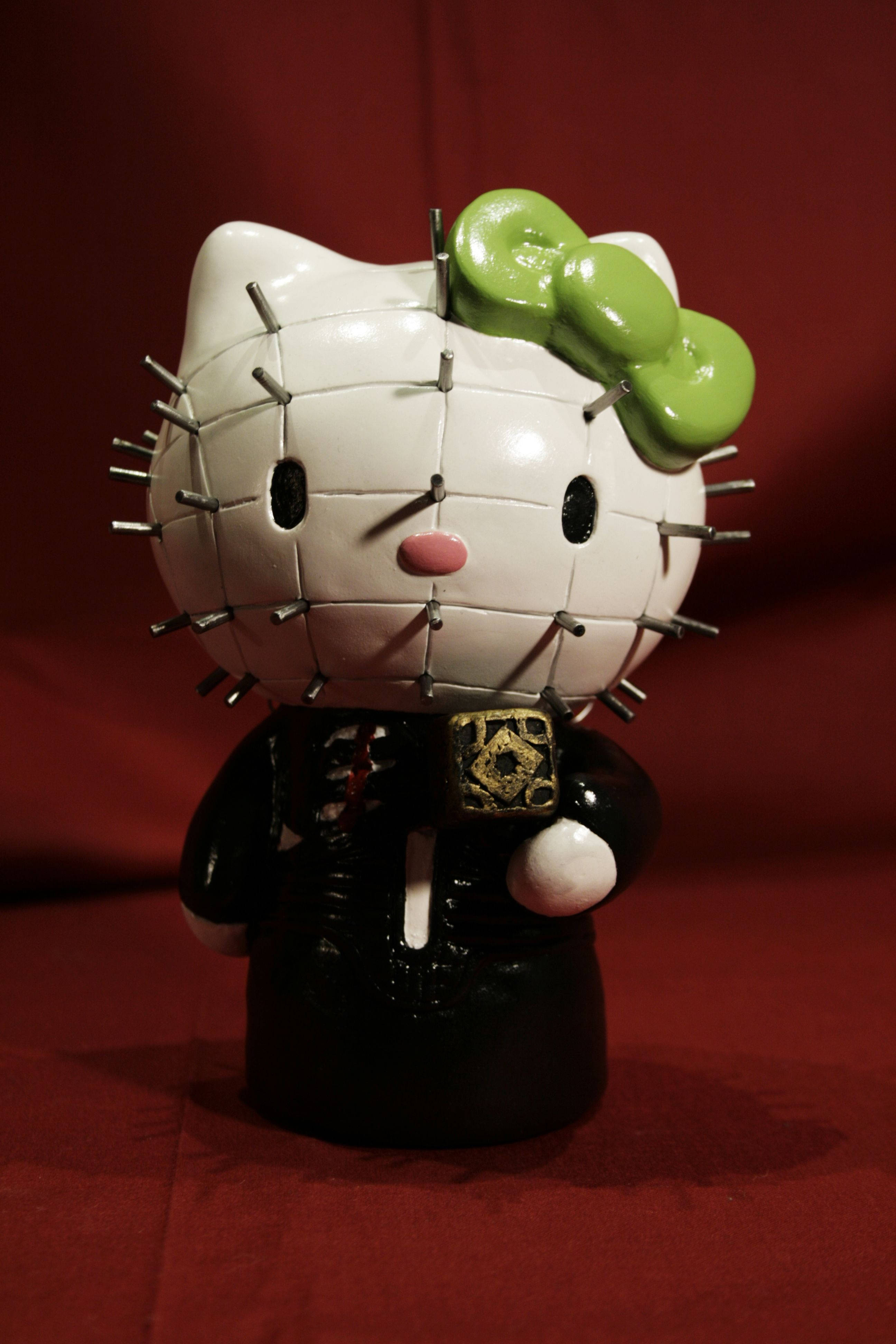 尖刺臉Hello Kitty萬聖節 壁紙 — 一個白色陶瓷Hello Kitty公仔，頭上同身上有綠色蝴蝶結同金屬針，企喺深紅色背景前面。