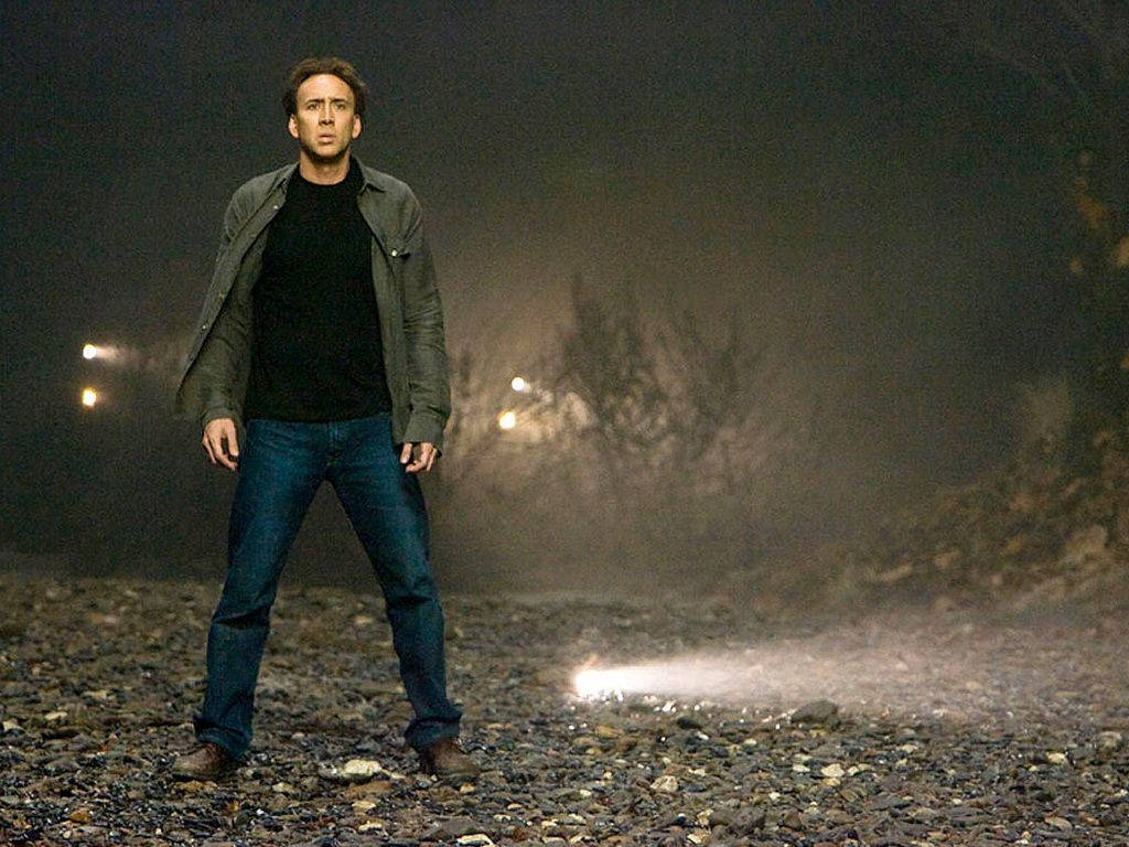 男人夜晚站在碎石路上，身後霧中有車頭燈。