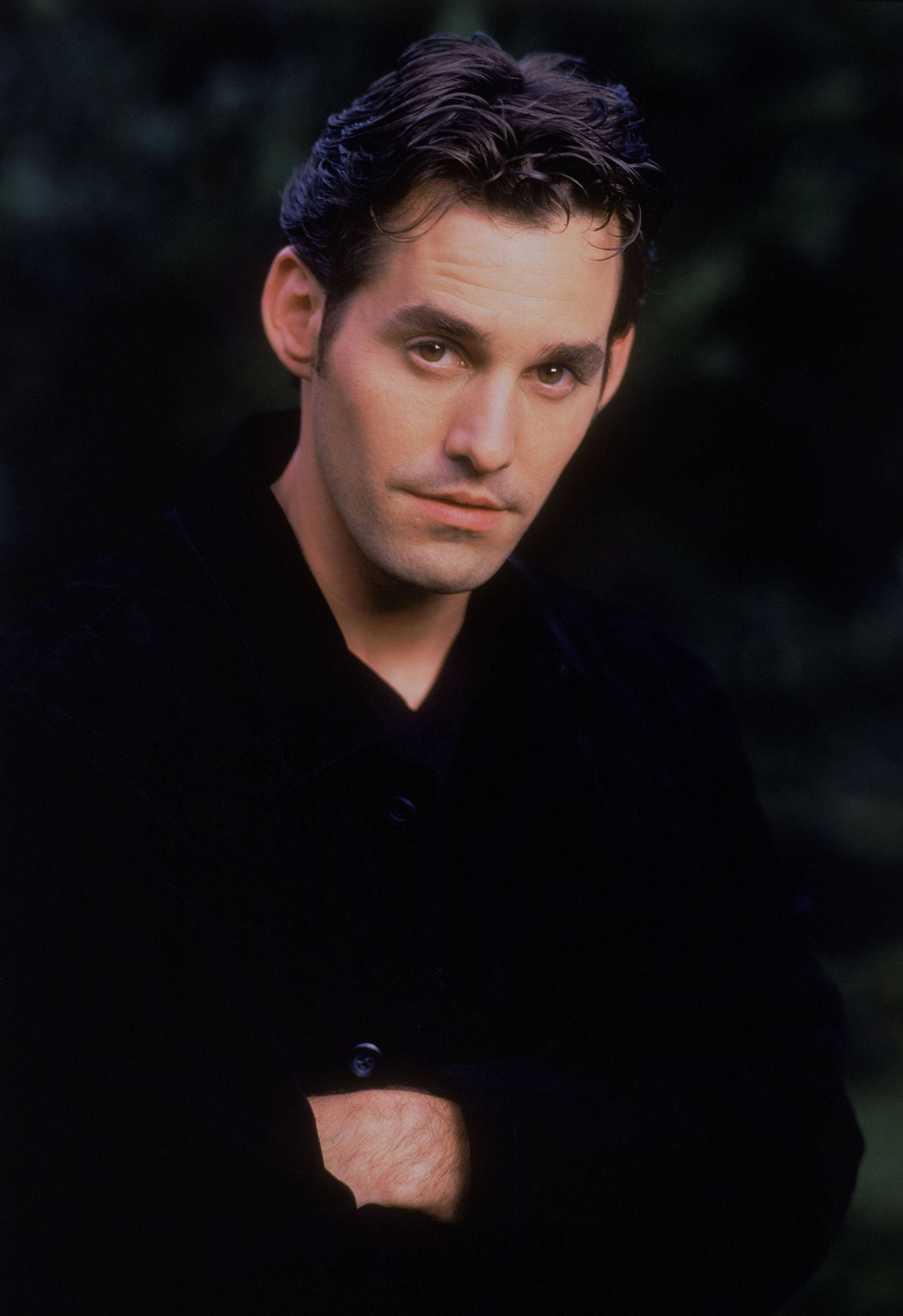 尼古拉斯·布雷登飾演Xander Harris 壁紙 — 一個穿著黑色外套的男人雙臂交叉站立，背景黑暗模糊，直視鏡頭，表情嚴肅。