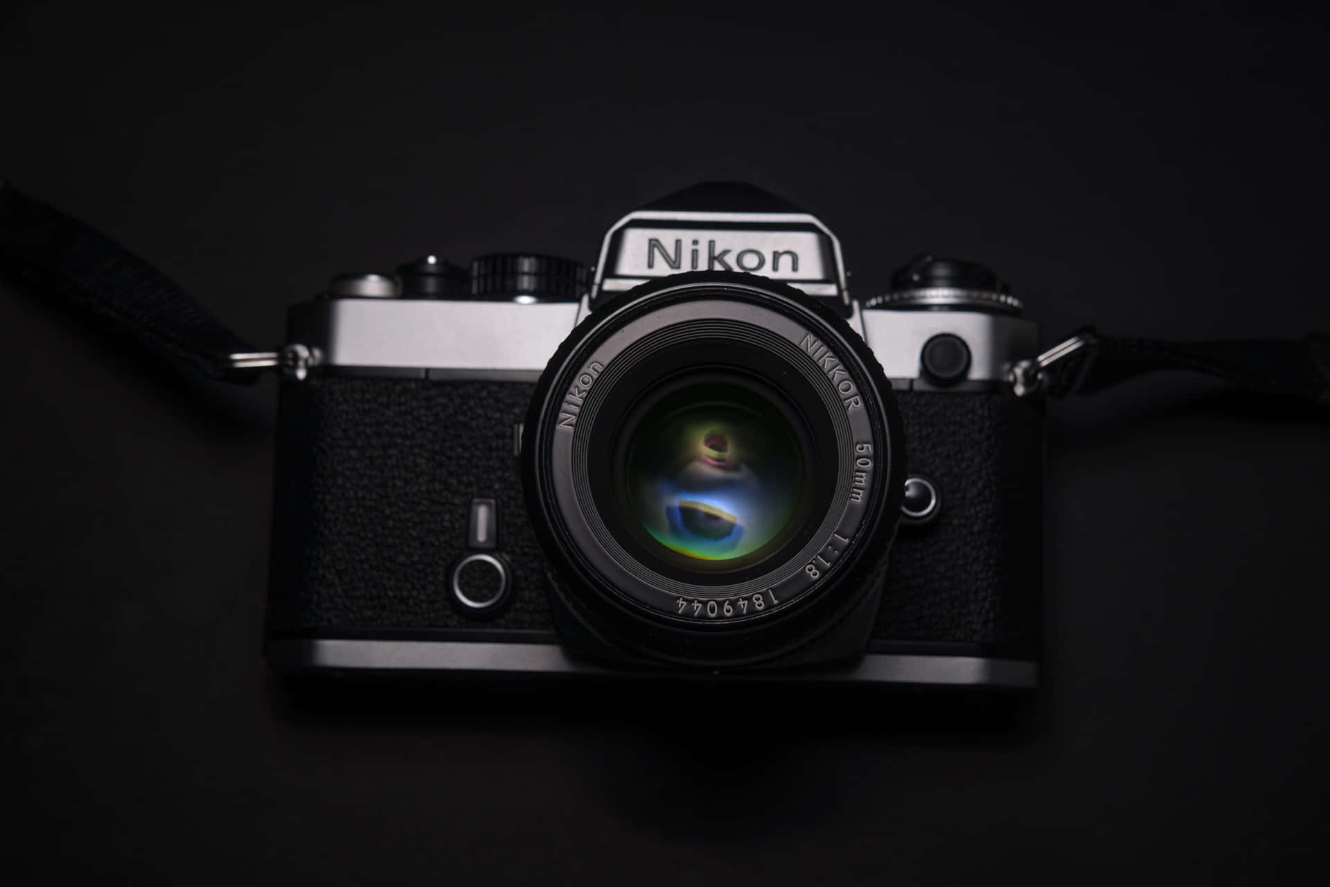 尼康F3單眼相機置於黑色背景 壁紙 — 一部復古 Nikon 菲林相機配備 50mm 鏡頭，放置在黑色表面上，鏡頭在昏暗的攝影棚中反射出鮮豔色彩。