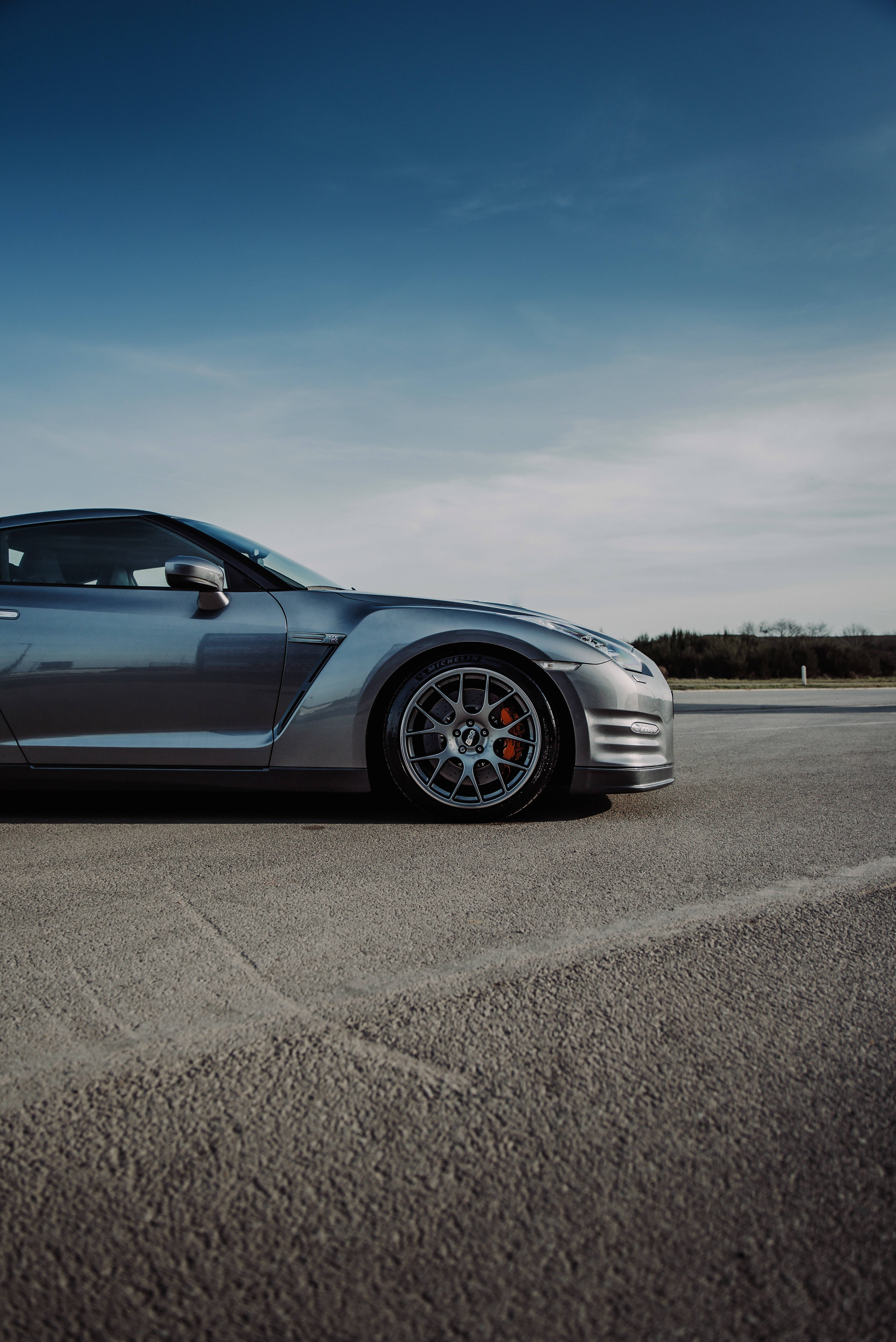 尼桑GT-R 定製側裙 壁紙 — 一架時尚的銀色跑車停泊在瀝青路上，頭頂是廣闊的藍天，點綴著縷縷薄雲。