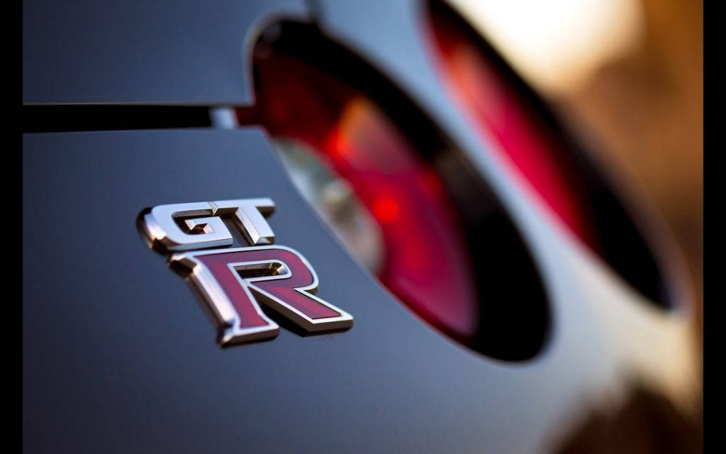 尼桑GT-R 標誌 壁紙 — 特寫鏡頭：在溫暖逆光下，光滑黑色車身上的閃亮GT-R標誌，紅色尾燈模糊。