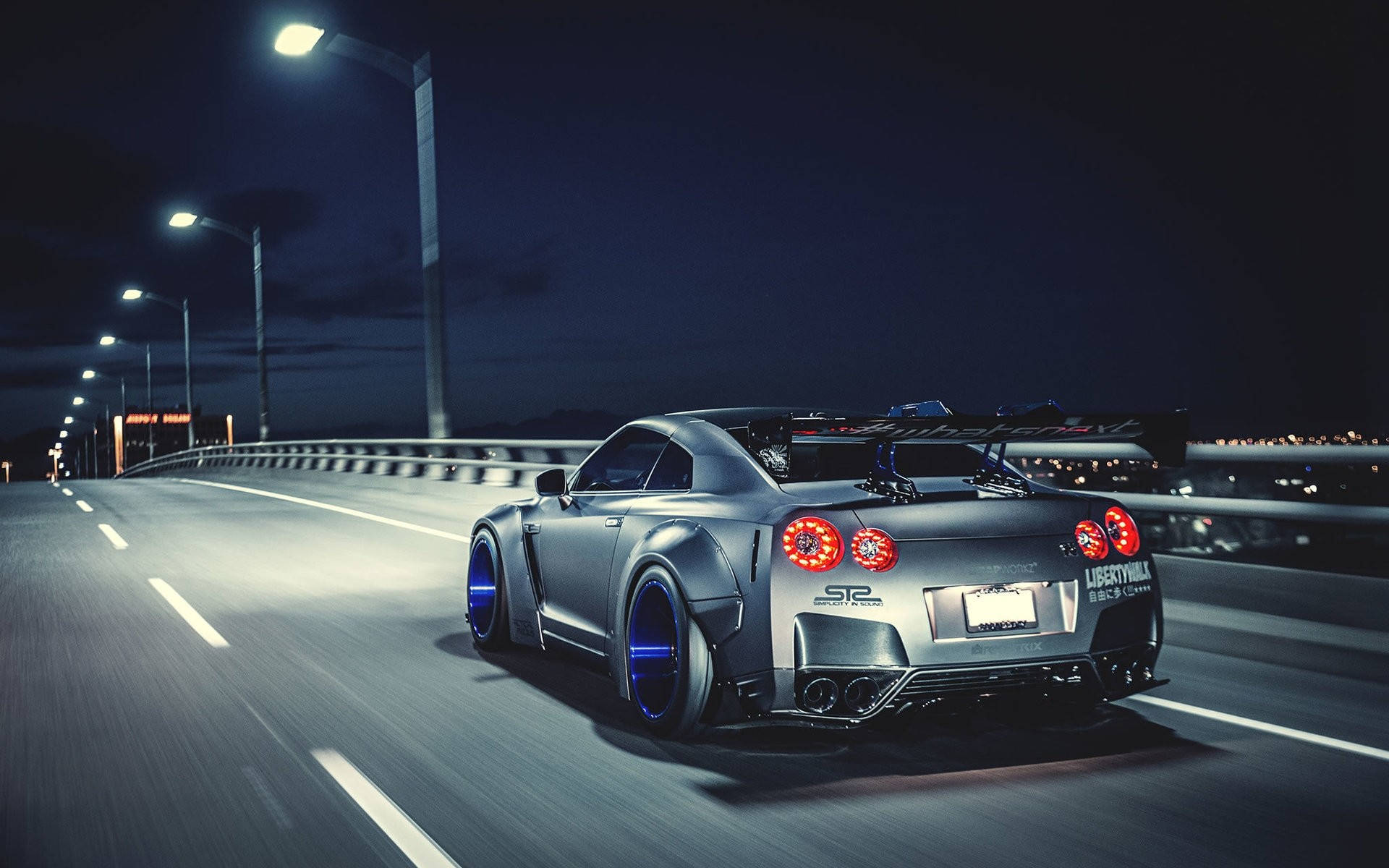 尼桑GT-R 紫色鍍鉻輪圈 壁紙 — 一輛銀色日產GT-R配備發光藍色輪圈，在夜間街燈下沿著昏暗的橋樑飛馳。