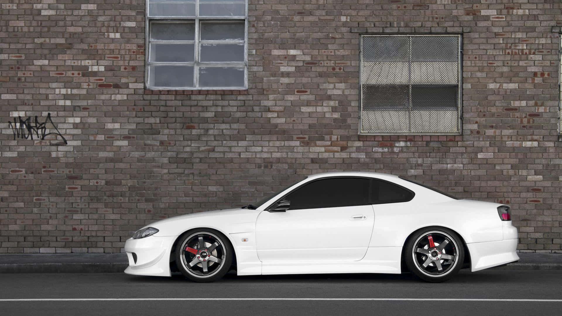 尼桑Silvia S15雙門跑車 壁紙 — 一輛時尚的白色跑車配備紅色制動卡鉗，停在一面帶有塗鴉和鐵窗的風化磚牆旁。