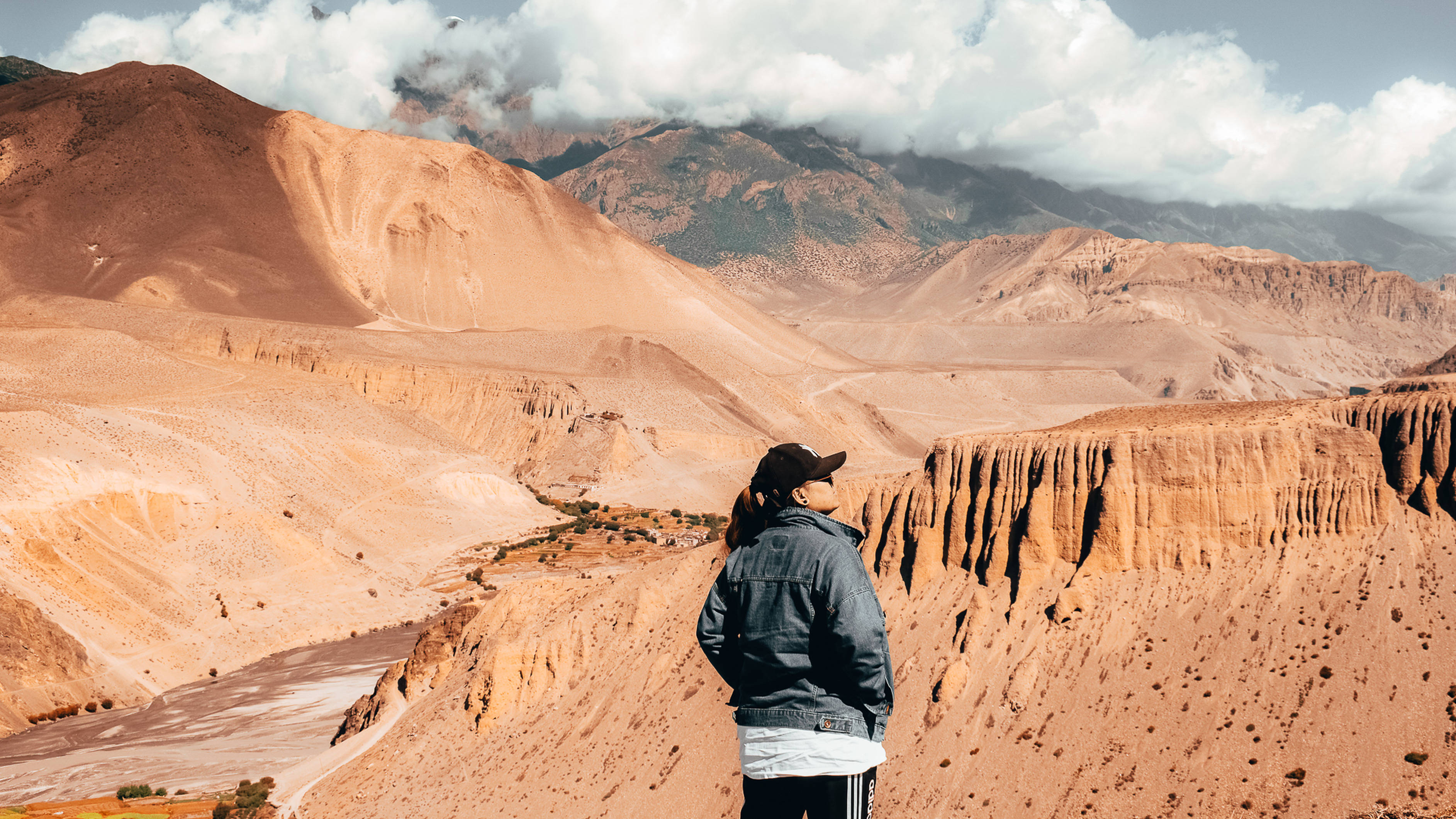 尼泊爾沙漠風光 壁紙 — 一位身穿牛仔褸的孤獨旅人站在崎嶇的峽谷邊緣，俯瞰著多雲天空下廣闊的橙紅色沙漠山谷。