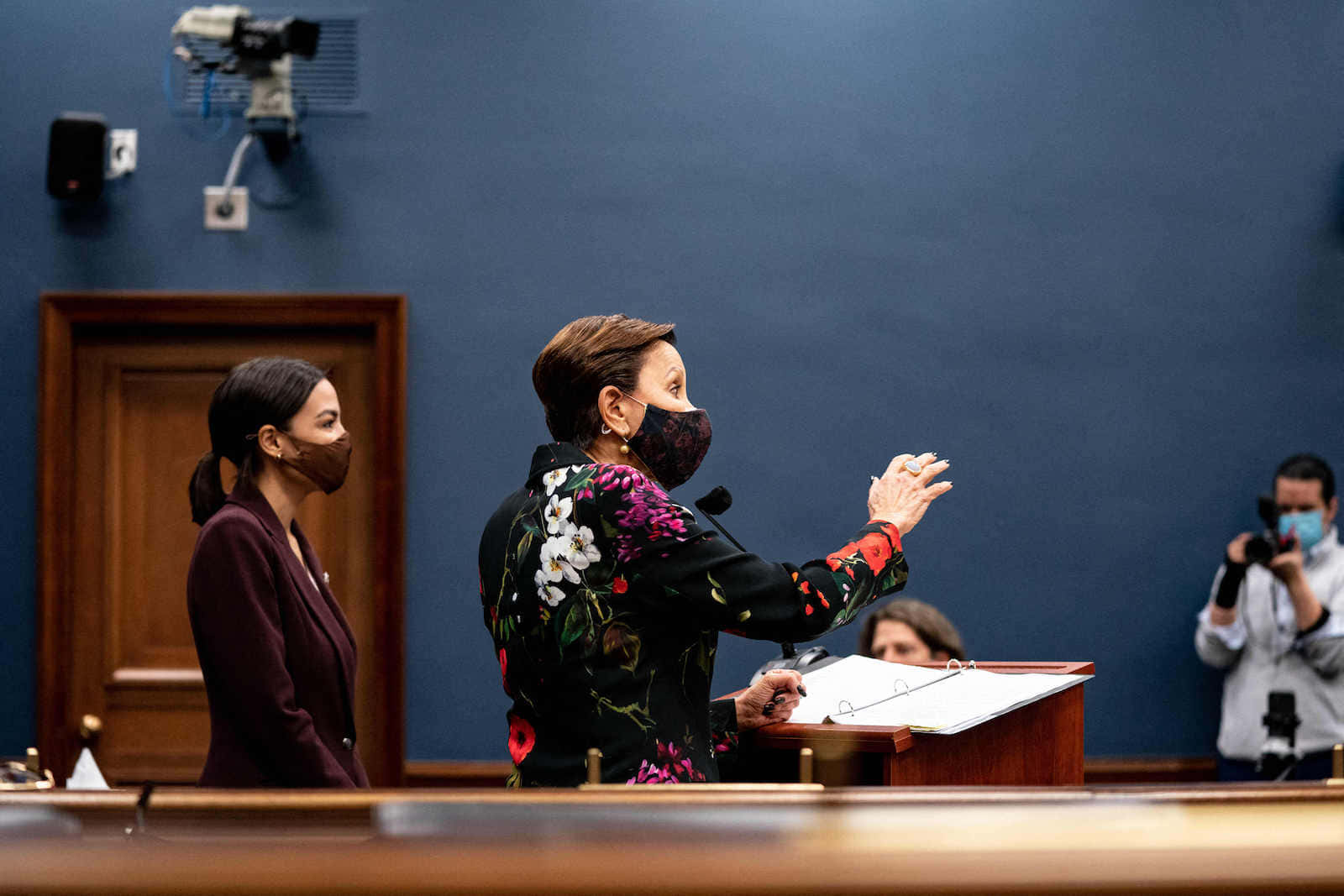 尼迪婭·維拉茲克斯戴黑色口罩 壁紙 — 一位身穿花卉圖案外套和戴口罩的女性在講台上強烈地做手勢，而另一位女性則在正式場合中站在附近。