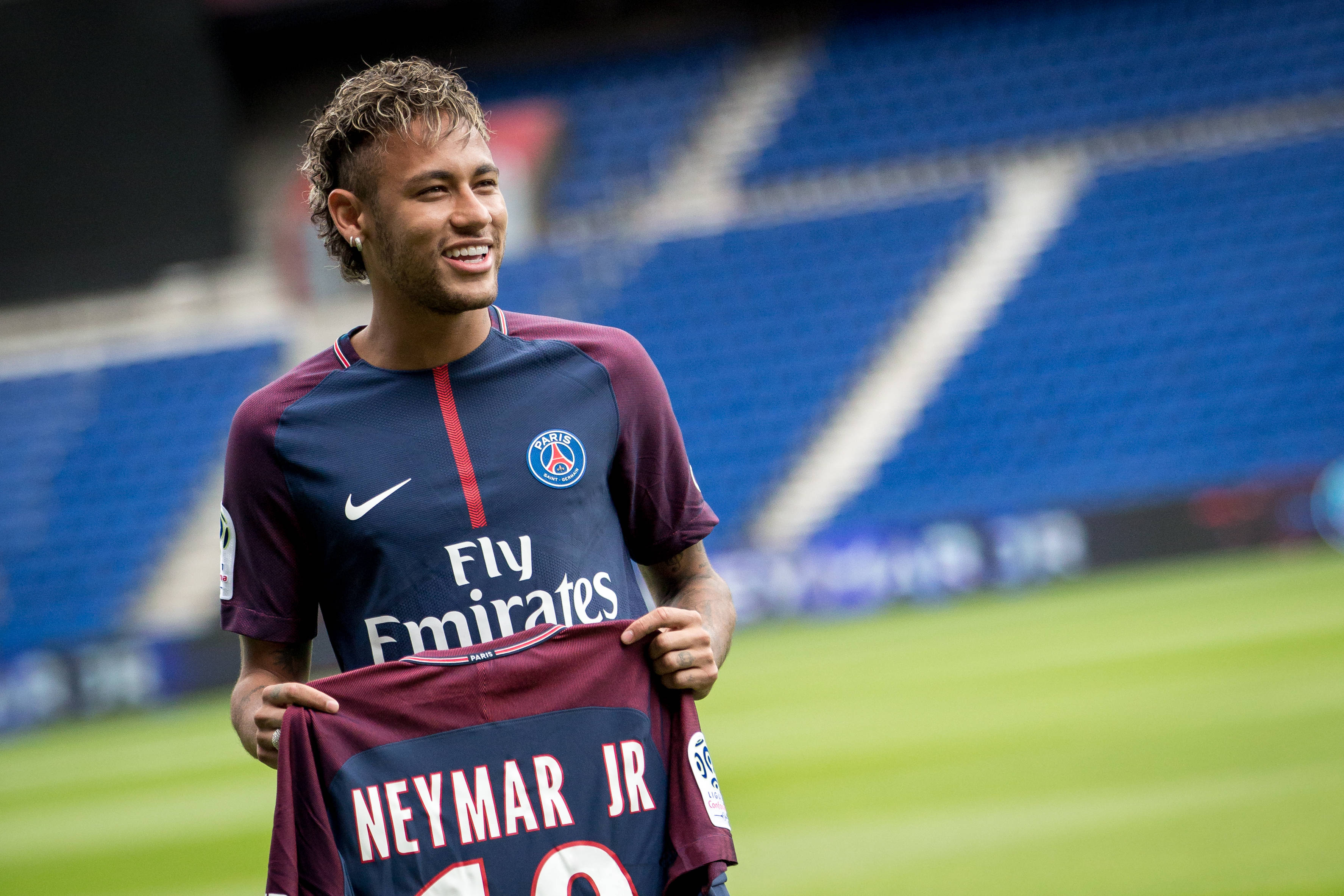 Brazil 桌布 壁紙 — 尼馬（Neymar Jr）在陽光普照的球場上，手持巴黎聖日耳曼球衣，露出燦爛的笑容。