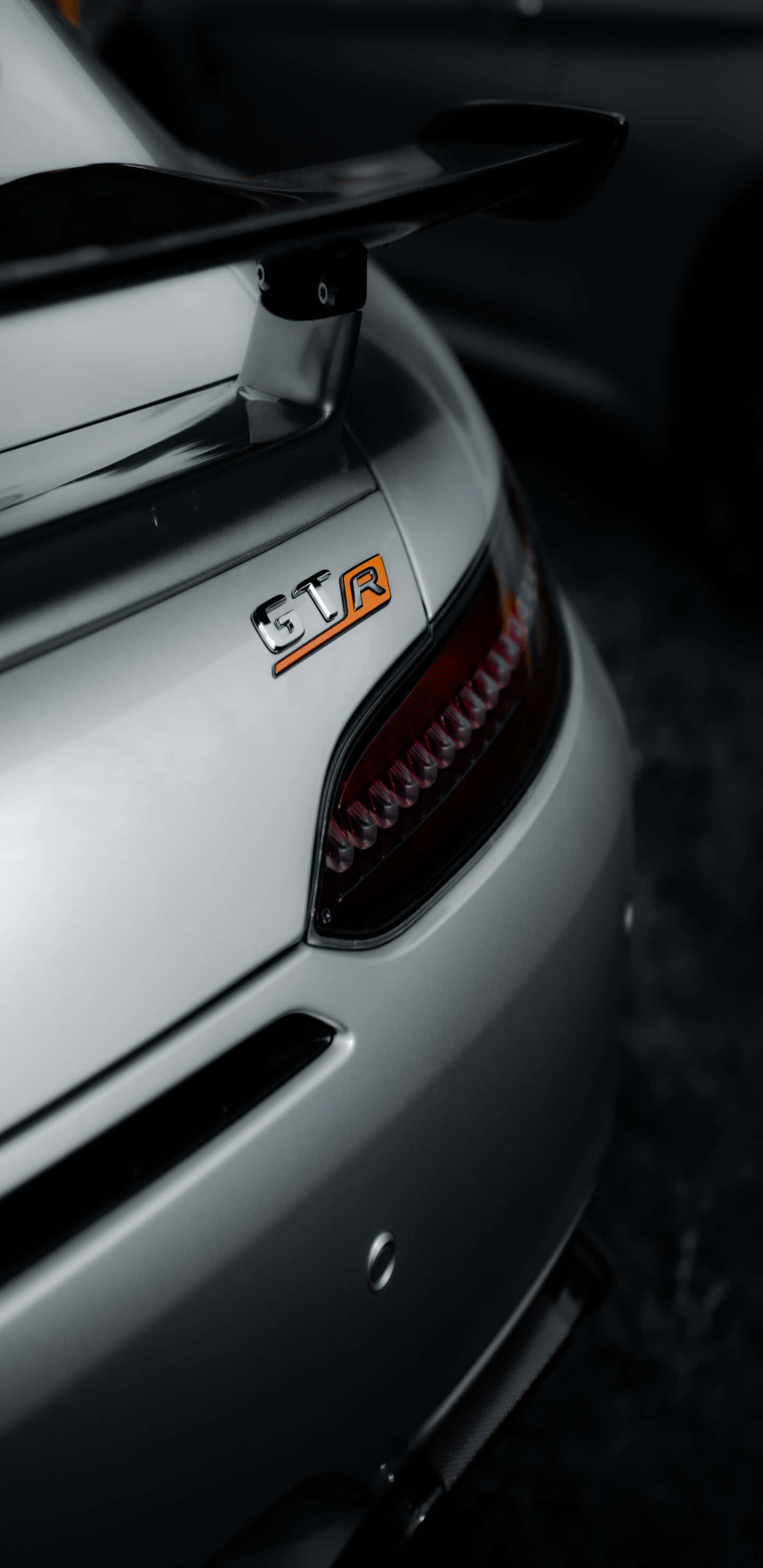 尾燈與梅賽德斯-奔馳GT-R標誌特寫 Pixel 3XL AMG GT-R背景 壁紙 — 銀色跑車尾部特寫，帶有橙色STR徽章和時尚尾燈，背景為深色。