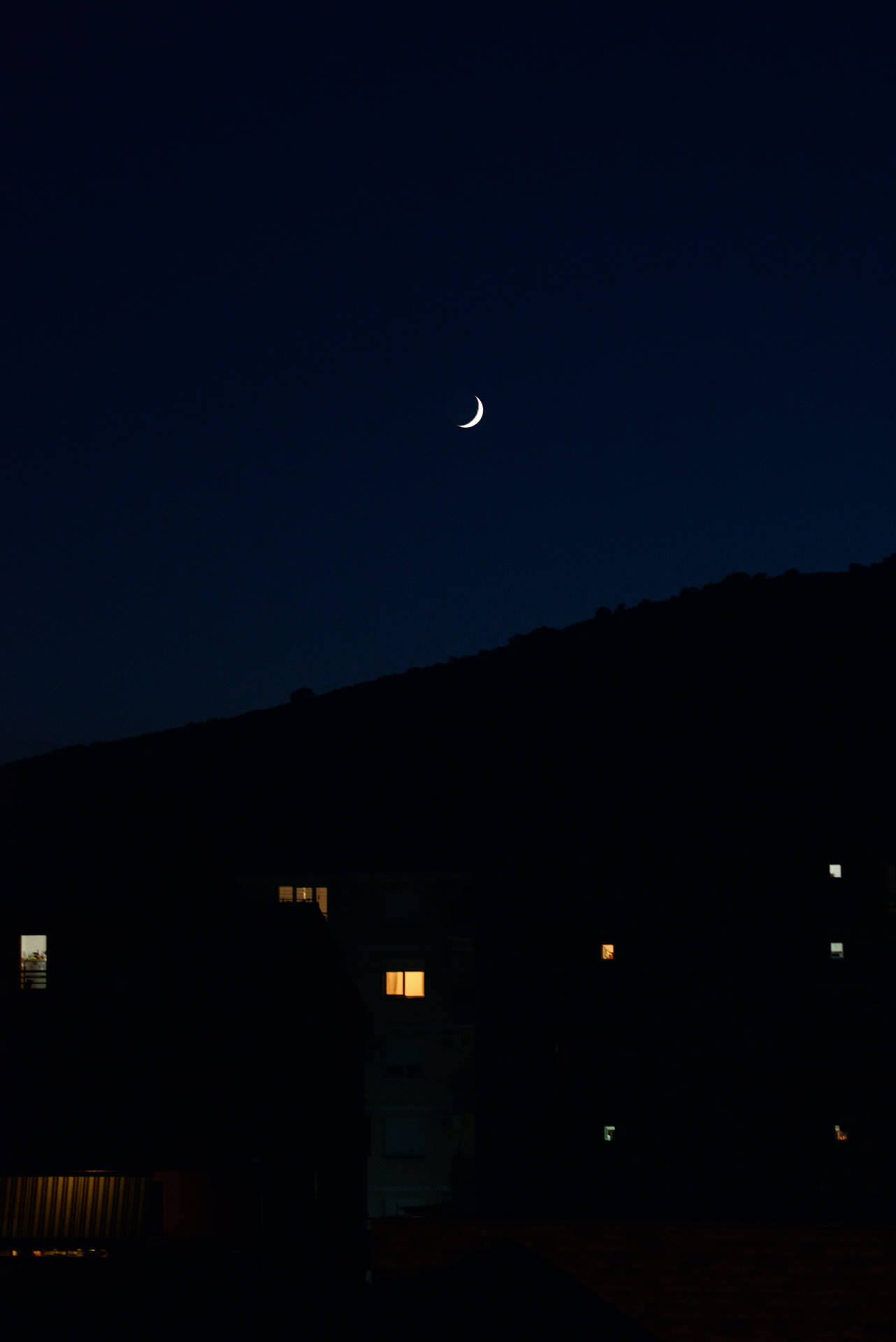 夜間建築物亮著窗戶的黑暗山丘上的月牙。