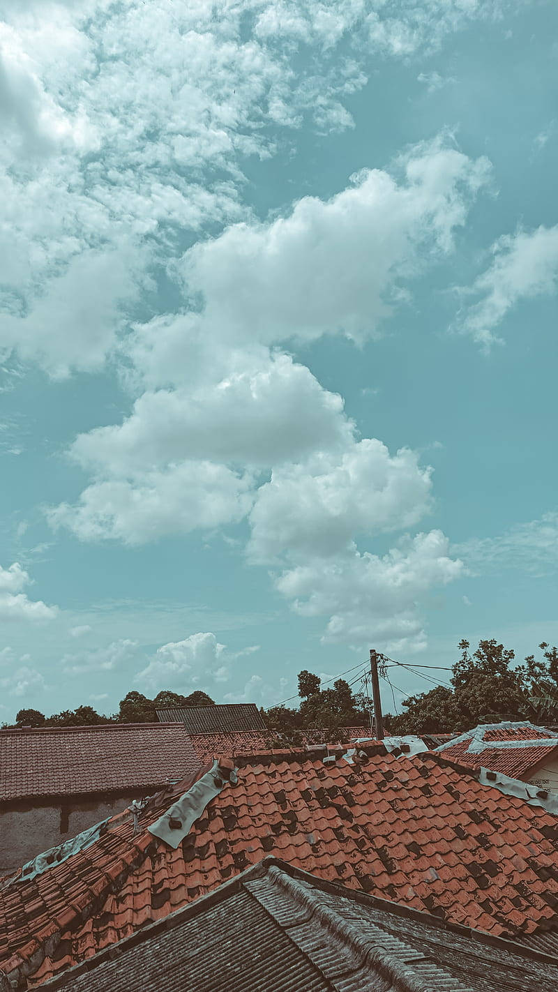 屋頂雲天手機壁紙 壁紙 — 紅色瓦片屋頂在廣闊的天空下延伸，天空充滿了蓬鬆的白雲和柔和的藍色色調。