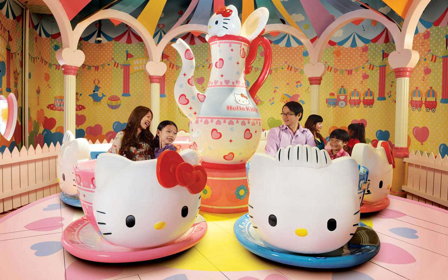 展現你對 Hello Kitty 的愛 壁紙 — 兒童和成人乘坐巨大的Hello Kitty茶杯遊樂設施，在色彩繽紛、夢幻的主題公園景點中。