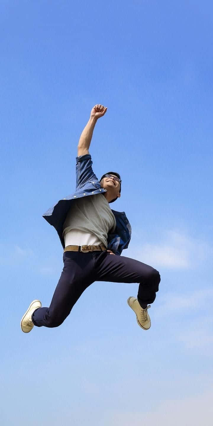 展現力量，#跳得更高 壁紙 — 一個快樂的男人在晴朗的藍天下高舉拳頭，高高躍起，充滿勝利感。