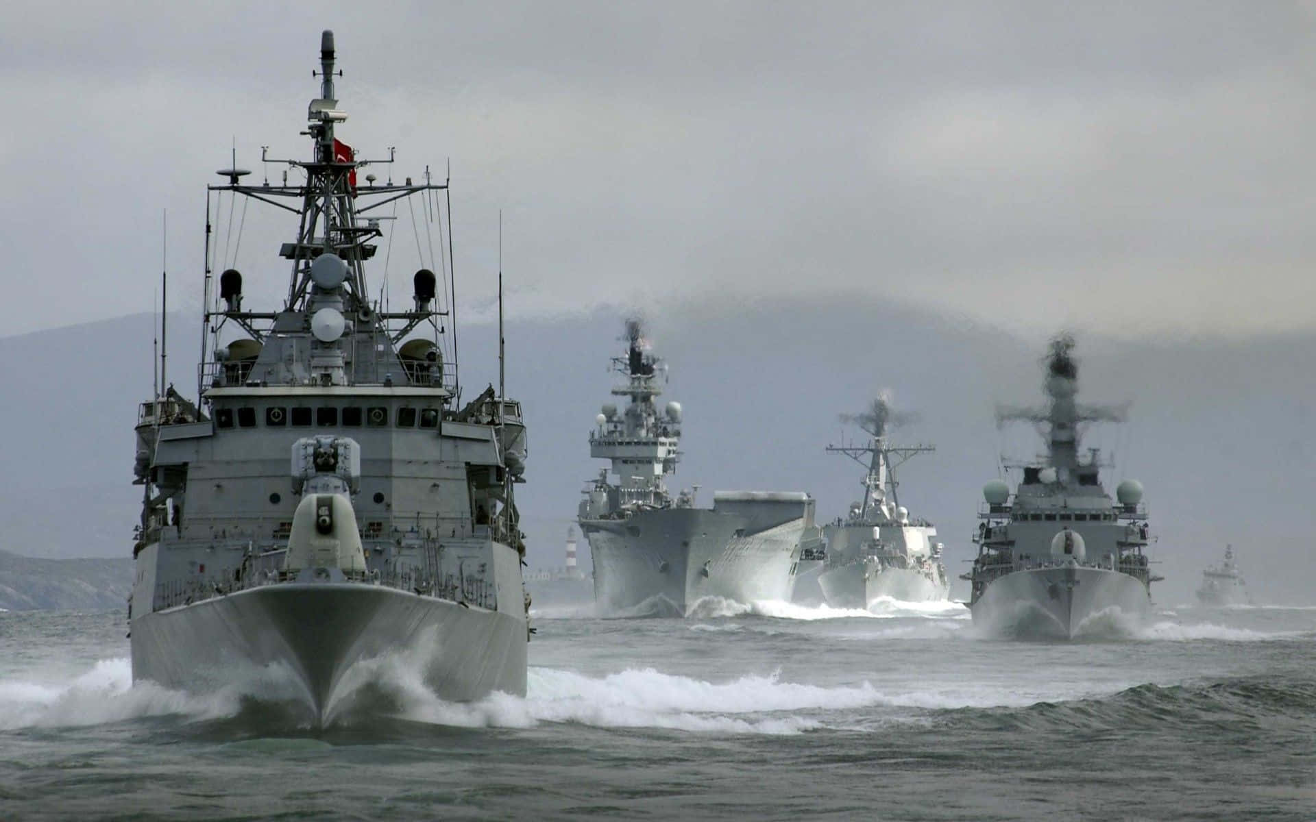 展現您的海軍榮譽！ 壁紙 — 一支灰色軍艦艦隊喺多雲嘅天空下，喺遠處群山嘅背景下，破浪前行。