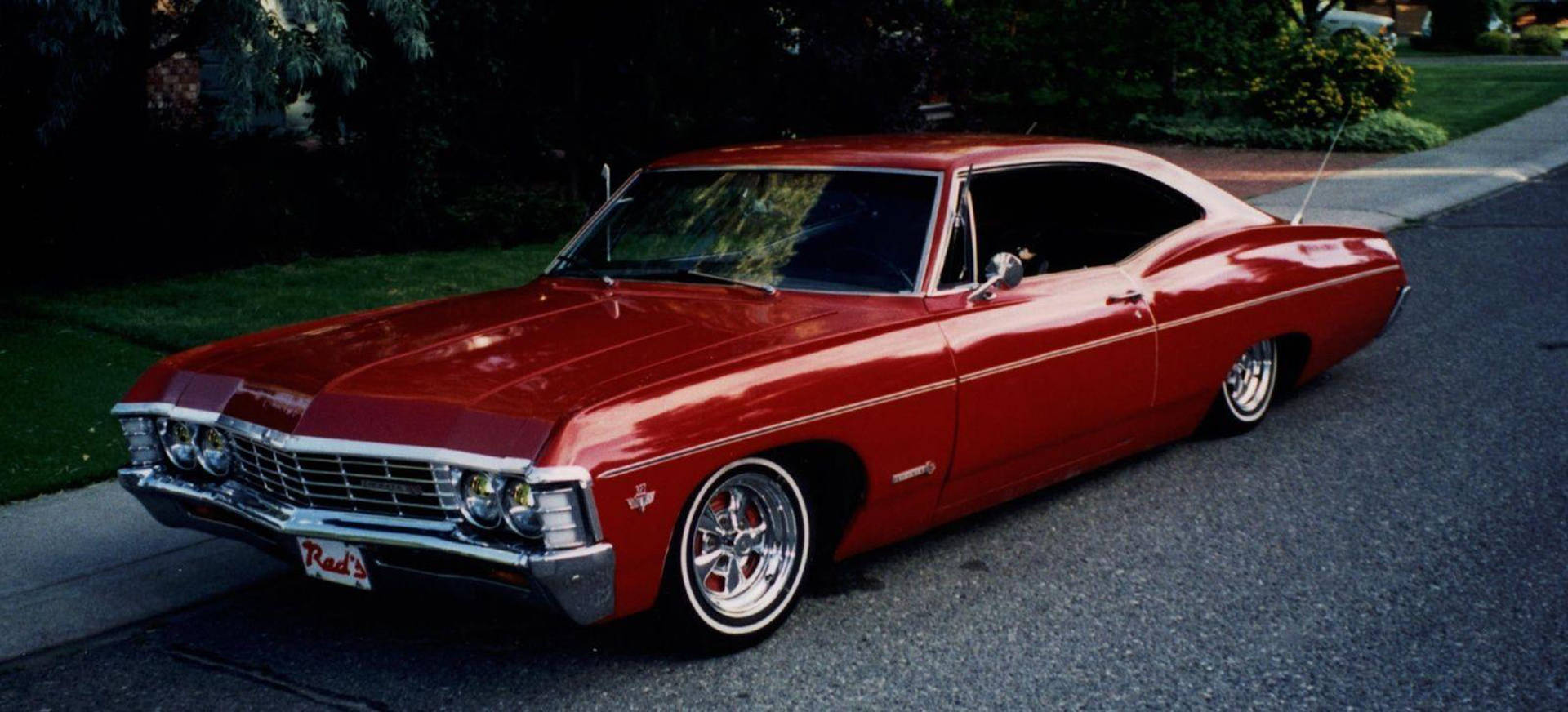 展現經典美學的 1967 年紅色雪佛蘭 Impala 壁紙 — 一架光面紅色經典肌肉車，帶有鍍鉻細節，停泊在黃昏的郊區街道上。