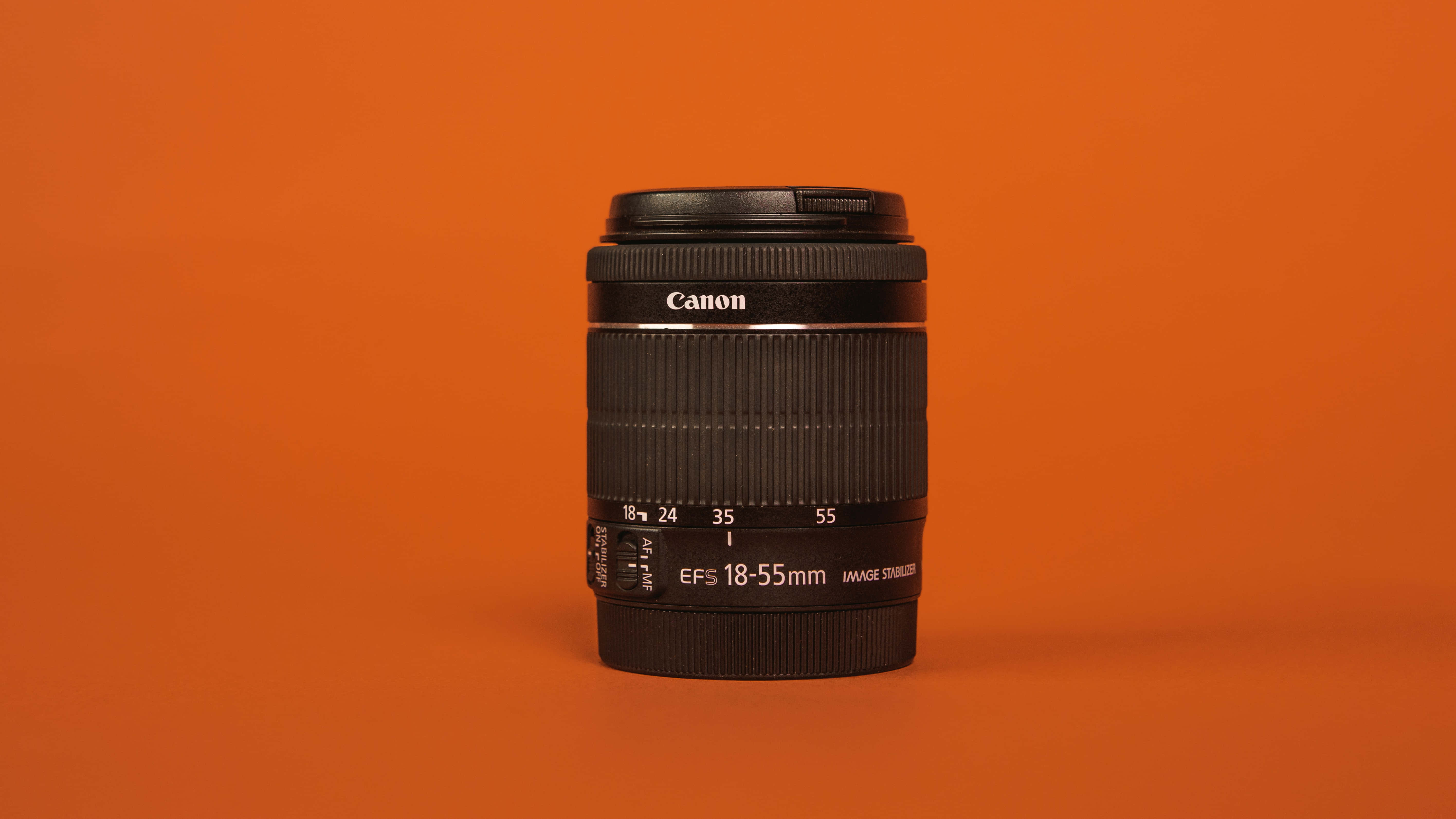 山丘地區令人著迷的風景 壁紙 — 一個黑色Canon EF-S 18-55mm相機鏡頭，居中放置在純色溫暖橙色背景上。