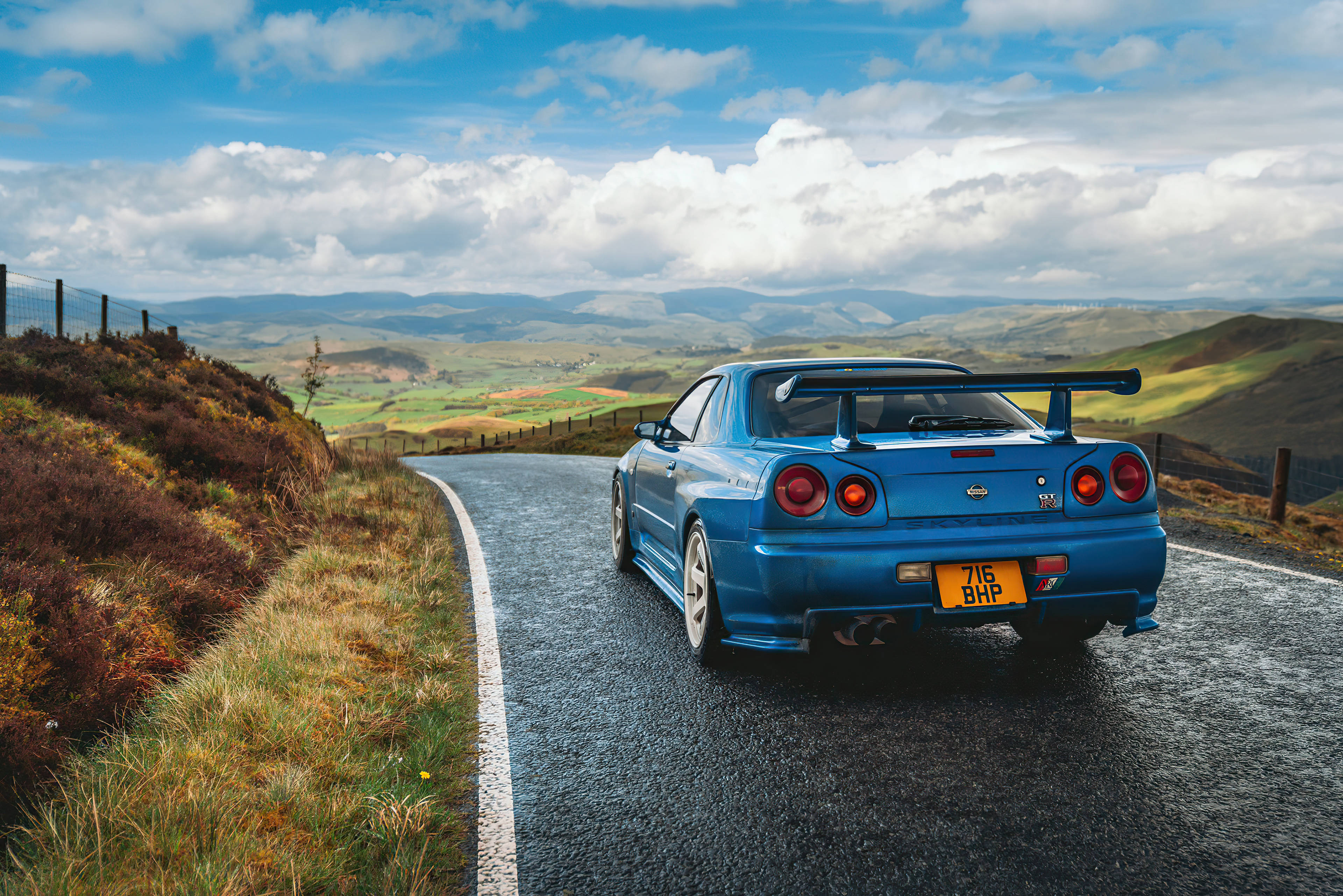 山丘路旁的藍色 Nissan Skyline GTR R34 壁紙 — 一輛充滿活力的藍色日產R34 GT-R在戲劇性的多雲天空下沿著蜿蜒的山路飛馳。