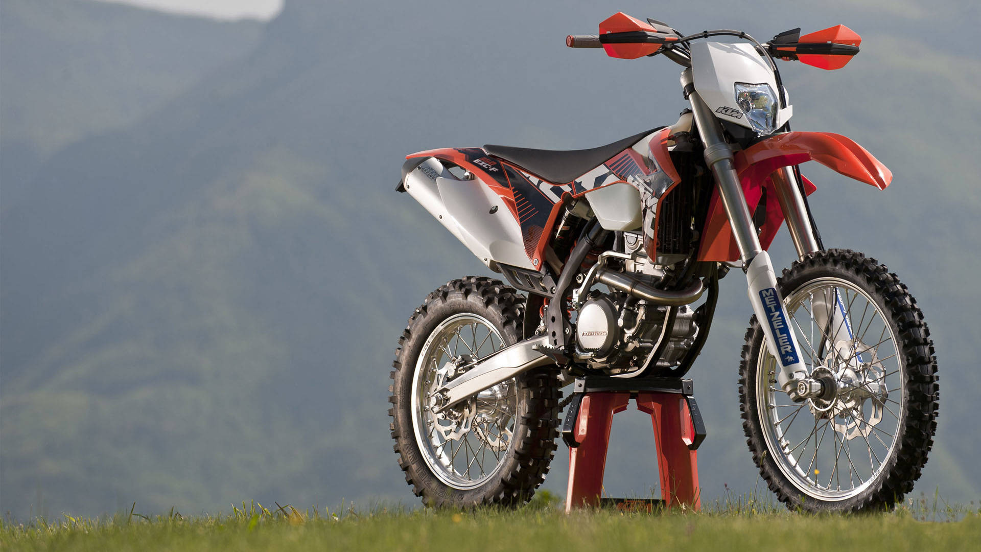 山區中的EXC-F 350 KTM越野車 壁紙 — 一架紅白越野電單車立於草地山丘上，背景是煙霧繚繞的綠色山脈。