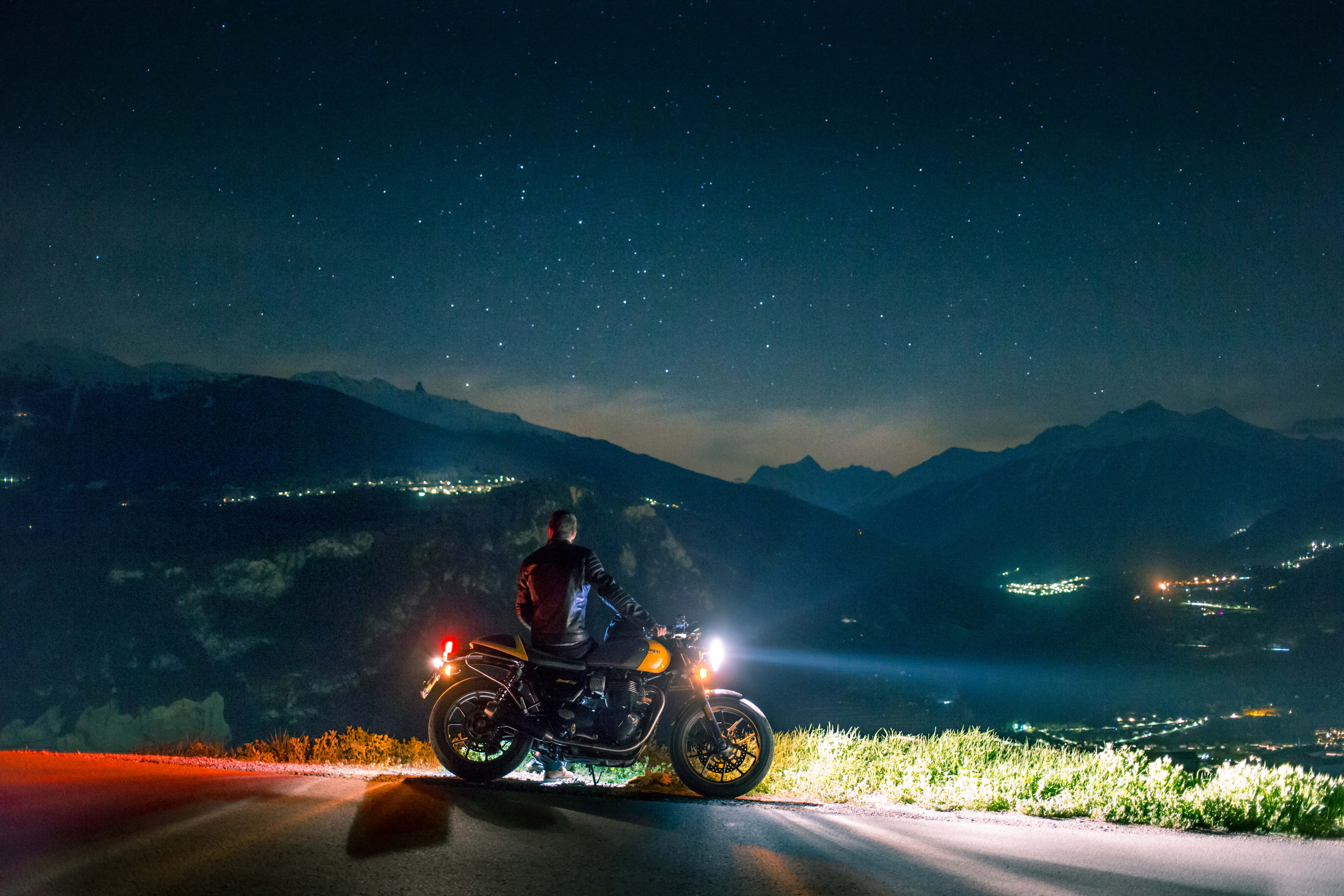 山區機車手谷地圖片 壁紙 — 一位孤獨的電單車手在星空下的夜晚山路上停下來，遠方的山谷燈光在下方閃爍。