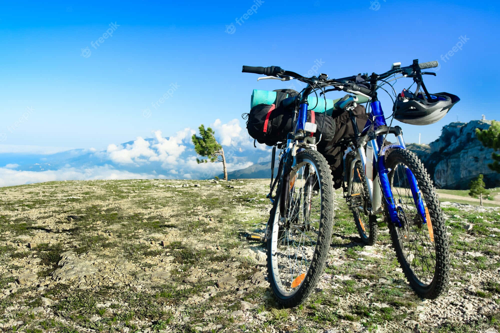 山地自行車與頭盔的運動風背景 壁紙 — 兩輛藍色山地自行車裝有裝備包，停放在岩石山頂上，俯瞰雲海和遠山，天空清澈湛藍。