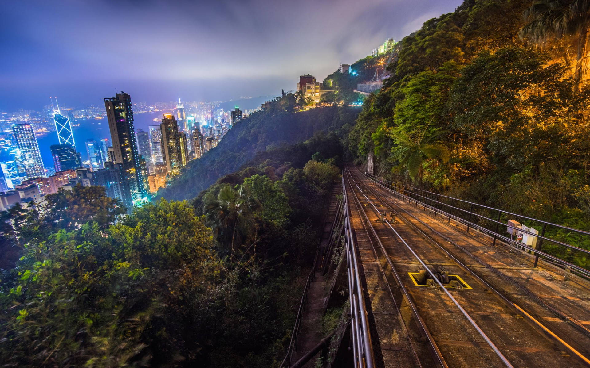 山坡鐵軌風景 壁紙 — 從蜿蜒穿過茂密綠意的陡峭山坡電車軌道上，欣賞香港璀璨的夜景天際線。