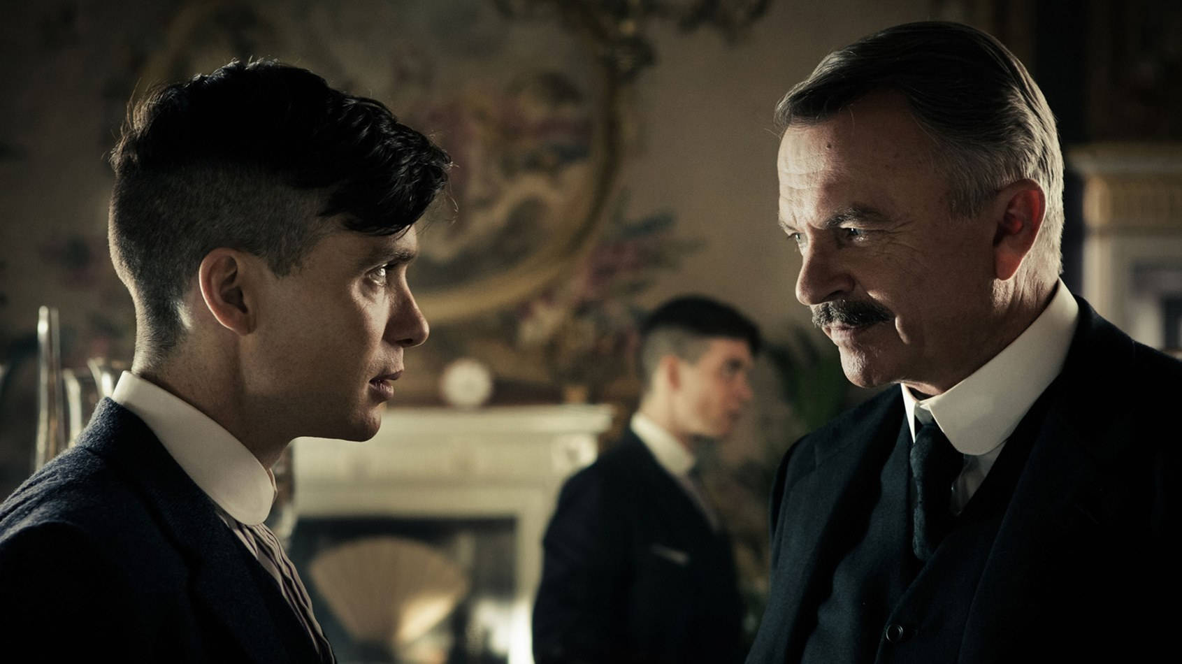 山姆·尼爾《Peaky Blinders》劇集劇照 壁紙 — 兩名穿著復古西裝的男子在一個光線昏暗、裝飾華麗的房間裡對峙，房間裡有壁爐和花卉壁紙。