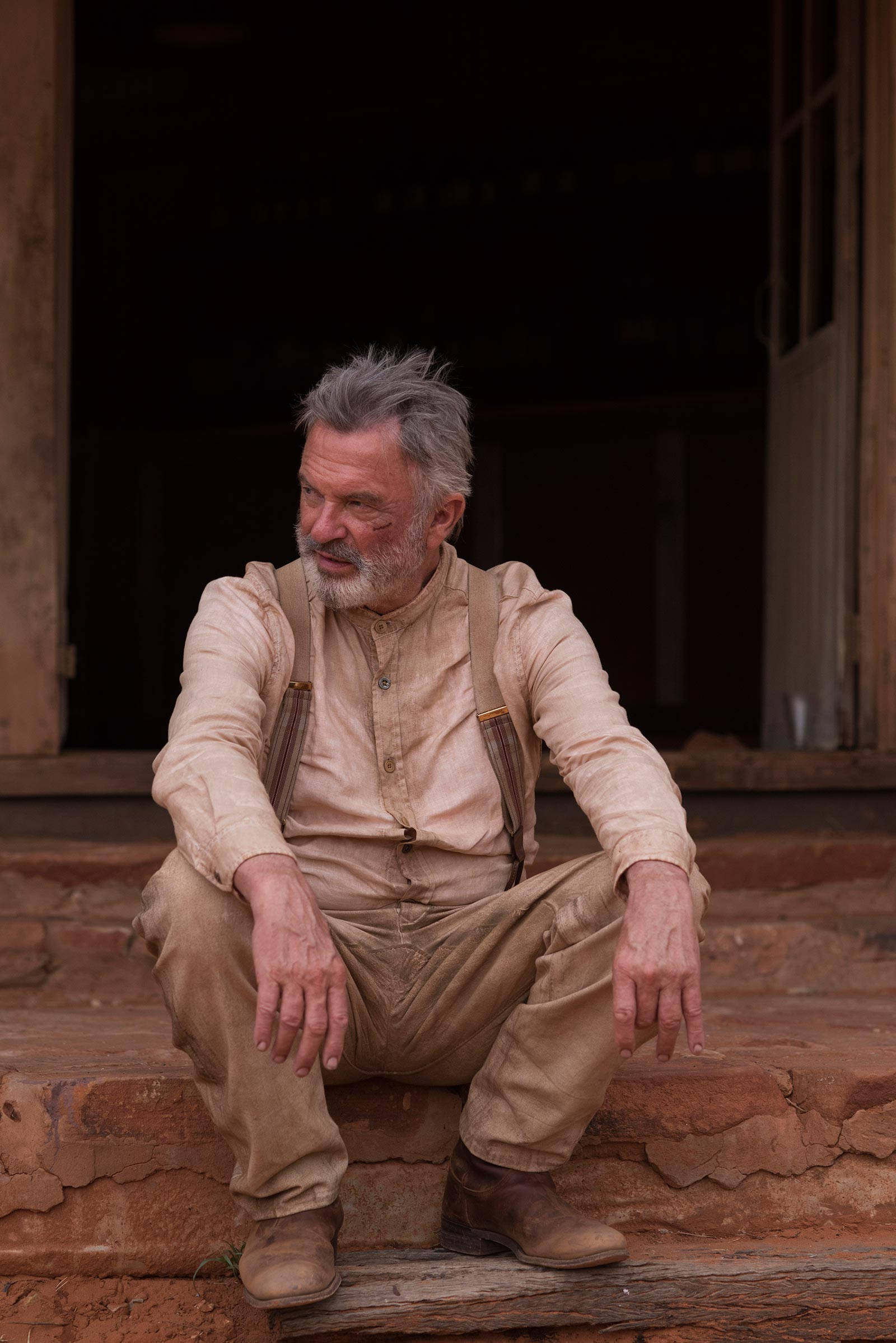 山姆·尼爾《Sweet Country》電影劇照 壁紙 — 一位灰白頭髮和鬍鬚的老年男子疲憊地坐在鄉村木門外風化的紅色石階上。