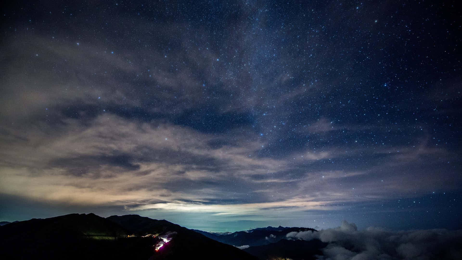 山巒上空的雲與繁星 壁紙 — 一個令人嘆為觀止的夜空，佈滿星星和縷縷雲彩，俯瞰著山脊，遠處的燈光蜿蜒穿過山谷。