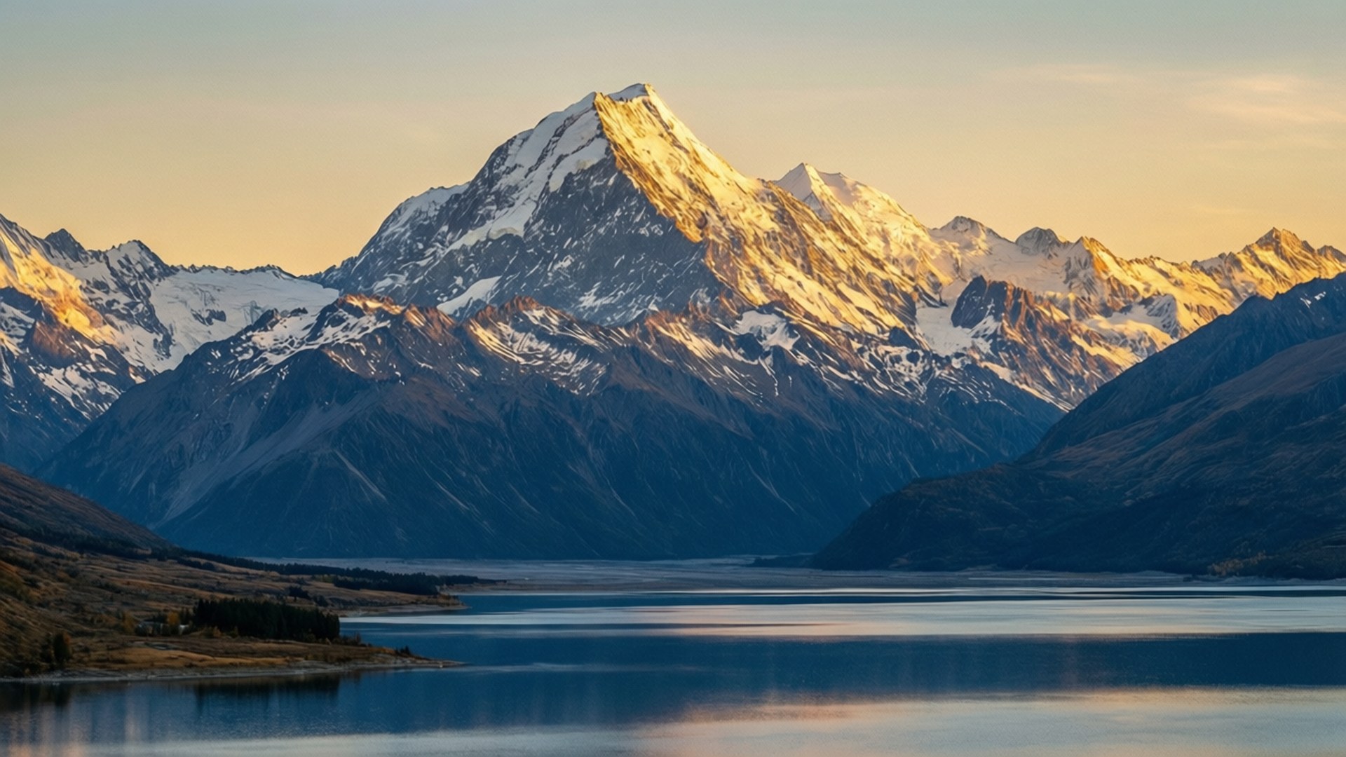 山巒日出全景 壁紙 — 白雪覆蓋的山峰在日出時金光閃閃，映照在寧靜阿爾卑斯山谷中平靜的湖面上。