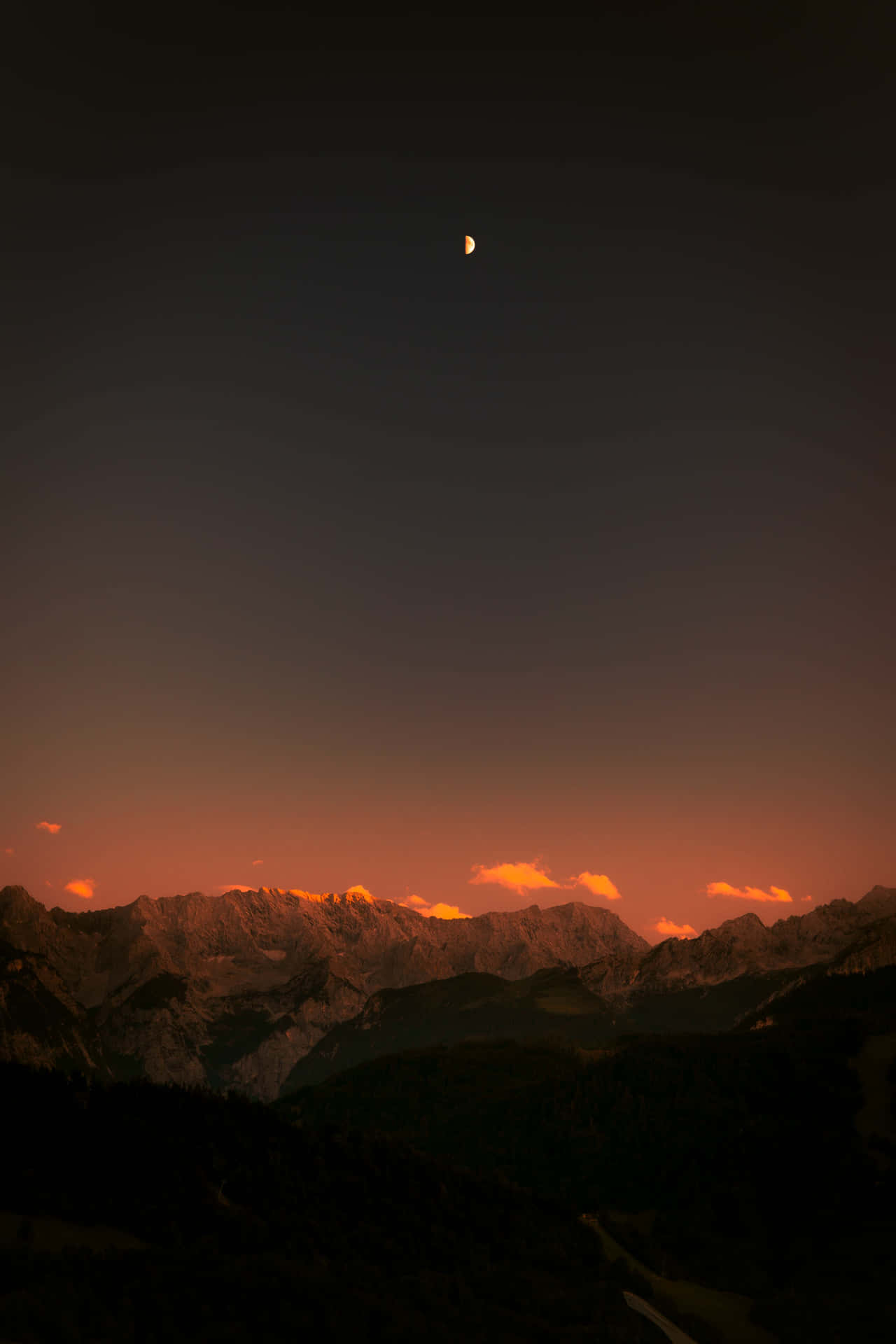 山巒日落月空 壁紙 — 一彎新月懸掛在鋸齒狀的山峰之上，山峰閃耀著熾熱的日落色調。