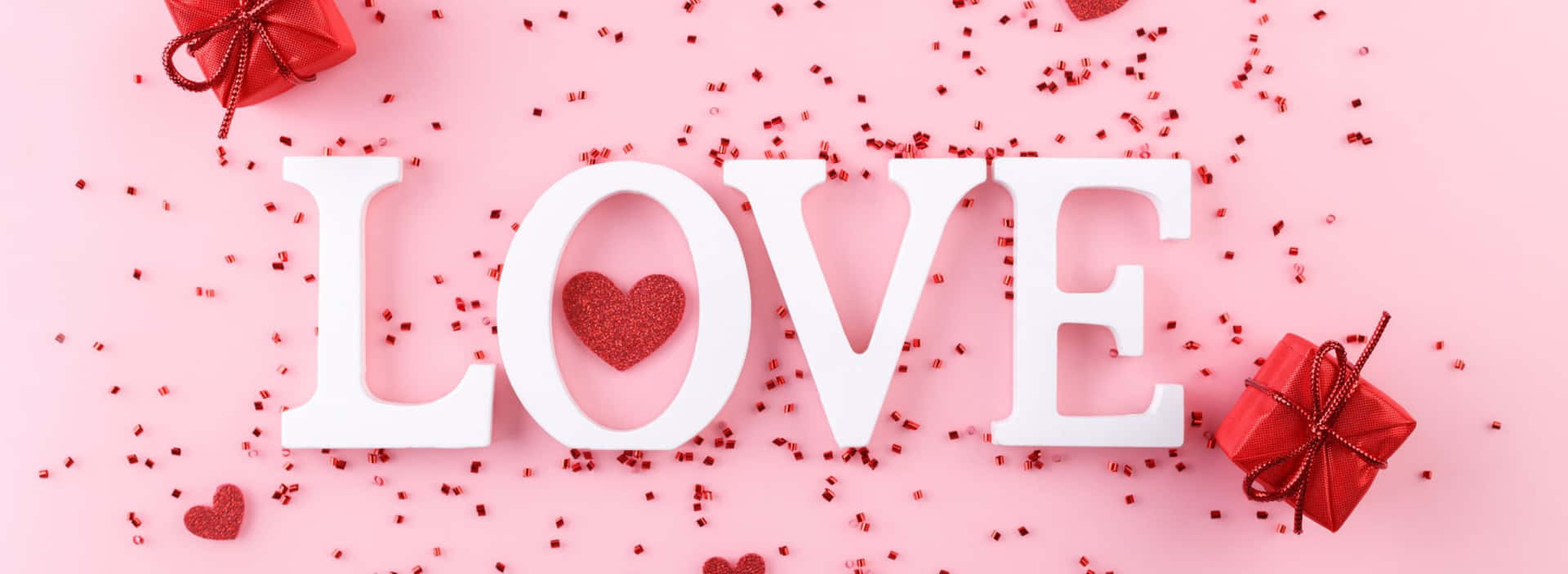 白色 LOVE 字母，帶有心形和紅色禮物，背景為粉紅色五彩紙屑。