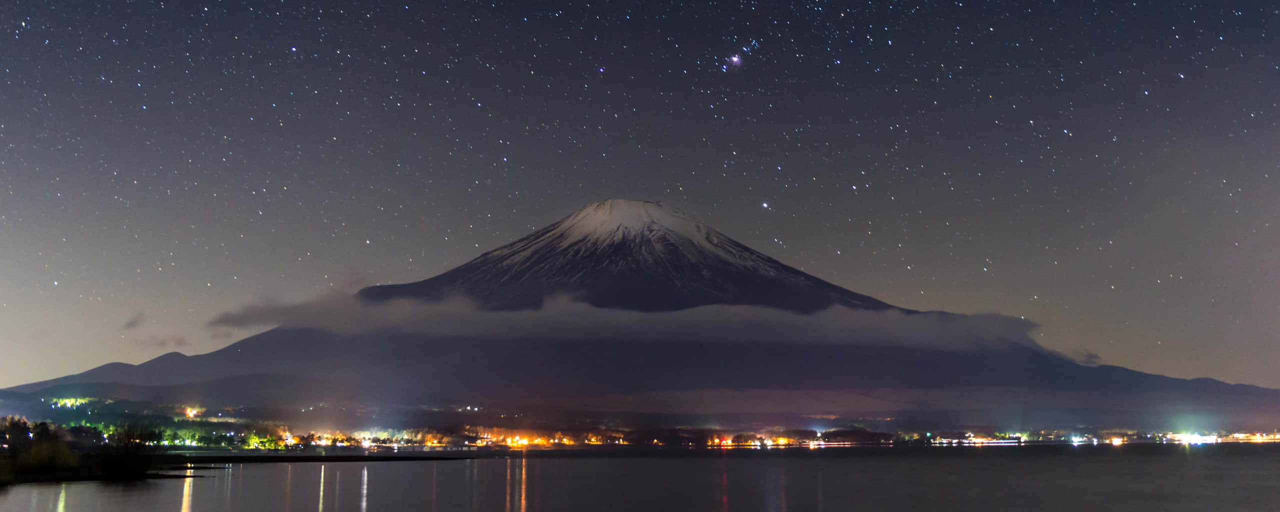 山巒超高清雙螢幕壁紙 壁紙 — 白雪覆蓋的富士山在繁星點點的夜空下雄偉聳立，城市燈光倒映在下方平靜的湖面上。
