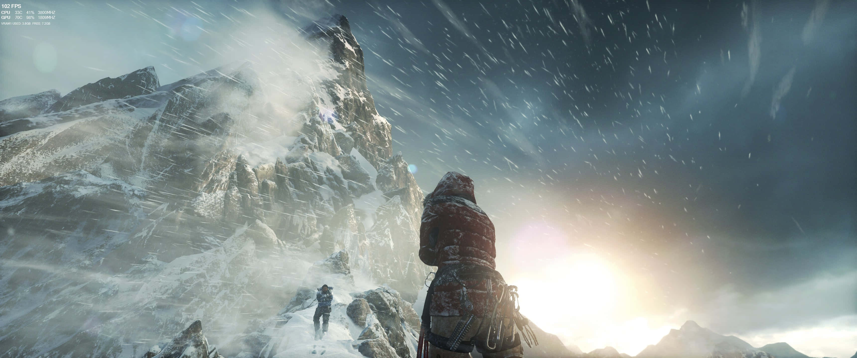 山巔 3440x1440p《古墓麗影：崛起》背景 壁紙 — 一名孤獨的登山者身穿重型裝備，在明亮的陽光下，風雪交加，站在白雪覆蓋的山峰前。
