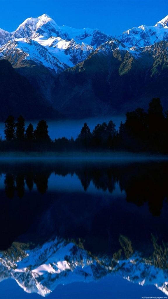 山影倒影自然 iPhone 背景 壁紙 — 積雪的山峰聳立在霧氣瀰漫的湖面上，在深藍色的暮色天空下映照出冰封的山巔。