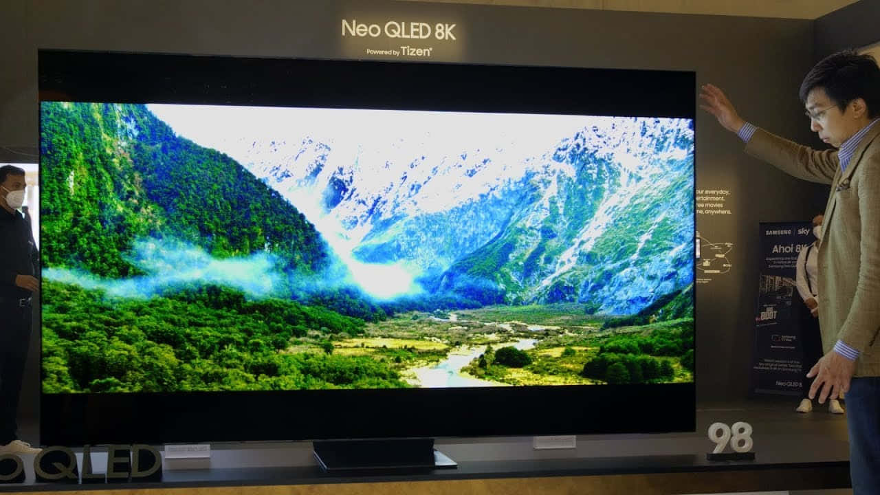 山景OLED電視畫面 壁紙 — 一名男子對著一部巨大的98吋Neo QLED 8K電視做手勢，電視上顯示著生動的山谷景色，有白雪覆蓋的山峰和茂密的綠色森林。